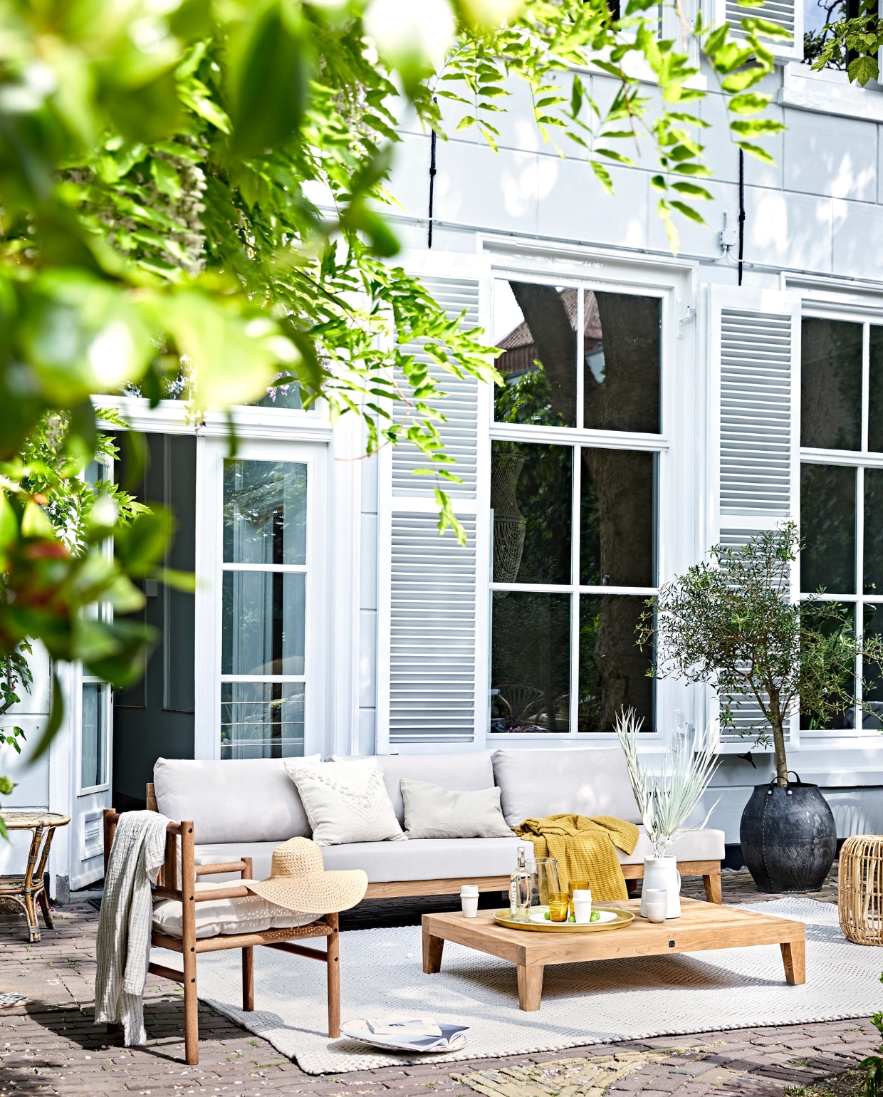 Buitentafel MBrace van Dedon 200x90 cm, houten stoelen Tibbo van Dedon (dacks.nl). Witte bistro klapstoelen van Fermob, witte armstoel Luxembourg van Fermob, Serviesgoed uit 12-delige dinner box Nordic Sand van Broste Copenhagen, schaal Nordic Sand van Broste Copenhagen ø 22,5 cm, Serax Surface longdrinkglas Serax Surface 4 stuks, linnen tafelkleed Gracie van Broste Copenhagen 160x300 cm (vtwonen.be). Gele servetten en wit linnen plaid (nordal.eu). Keramieken schaal op klapstoel, linnen lampionnen (sukha.nl). Ladder vanaf (com-anders.nl). Witte vaas (zarahome.com). Stonewashed bestekset Brick Lane 39-delig (luxerybynature.nl). Glazen kan (weldaad.com). Klapkruk van mangohout (hm.com). Okergeel kussen (madamstoltz.dk). vtwonen 07-2020 | styling Cleo Scheulderman | fotografie Jeroen van der Spek
