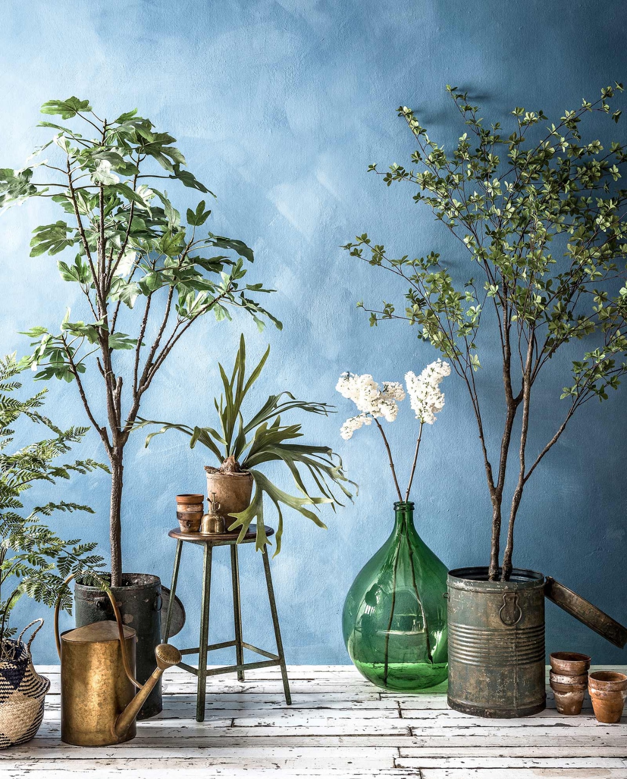 Muurverf in kleur Periwinkle van Bauwerk Color. Gevlochten mand met blauwe vlakken (). Kleine kunstplanten in pot, grote kunstplanten en -boompjes, groene metalen ton met deksel, houten kruk met metalen onderstel (). Messing gieter (). Oude terracotta bloempotten, witte seringen Messing plantenspuit (). Groene glazen flesvaas () vtwonen 2017-08 | fotografie Sjoerd Eickmans | styling Danielle Verheul