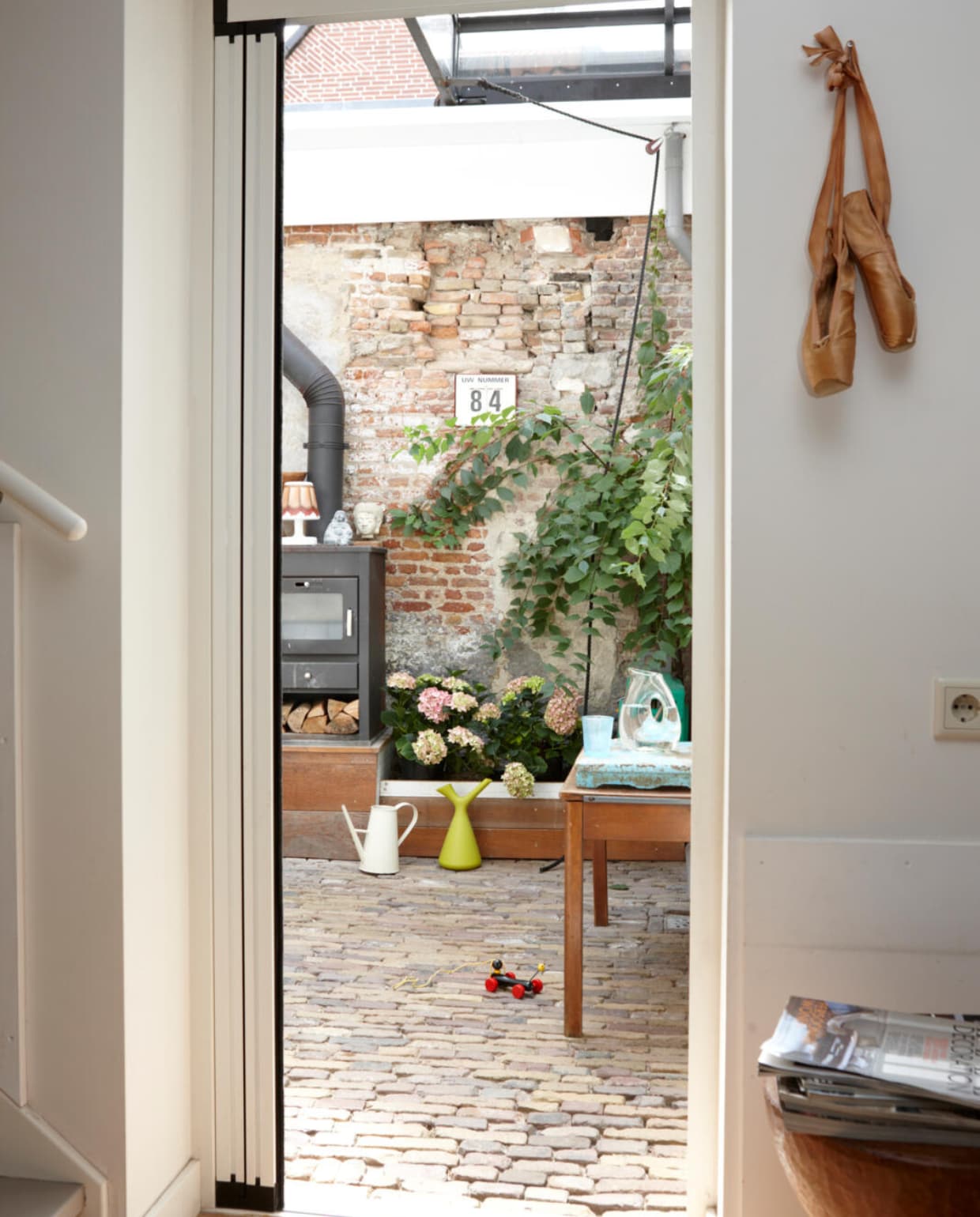 Doorkijkje naar de binnentuin, die afgesloten kan worden door middel van een glazen roldak. Een vriend heeft dit systeem met katrolmechanisme, eigenlijk een metalen frame met een glazen puntdak op wielen, gemaakt. Schuifdak van Heavy Decor. Gieter van Goods, karaf en glas van Pols Potten, lampje van Edison Fatboy. De boeddha en het beeldje op de houtkachel komen uit Indonesië. Het volgnummer-apparaat tikte Coen op een Belgisch marktje op de kop. Het oude lessenaartje een vondst van Marktplaats. vtwonen 03-2016 | styling Maya Burghouts | fotografie Simone de Geus