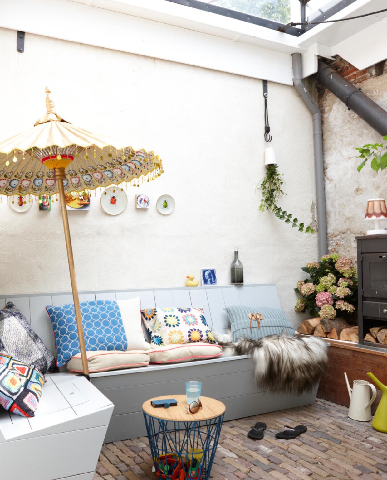 De knusse binnentuin waar het dak open en dicht kan. Ook hier een houten zitbank met veel opbergruimte, ontworpen en gemaakt door Coen en zijn vader. Parasol uit Indonesië, muurdecoratie van Wallware en Zolder op Stelten. Fles en glas van Pols Potten. Kussens, van links naar rechts: Granny van Snurk, een grijs kussen uit Marrakech, een met blauwe bollen van Design on Stock, nog een Granny van Snurk en een kussen van Knit Factory. De platte kussens komen van Leen Bakker. Mand en deksel van Ferm Living. Vacht van IJslands schaap en sky planter plantenhanger van Kavel84. vtwonen 03-2016 | styling Maya Burghouts | fotografie Simone de Geus