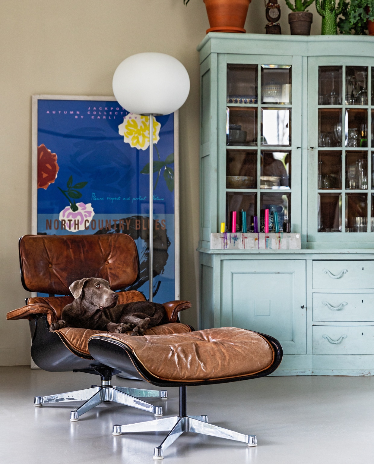 Eames Lounge Chair van Vitra met daarin hond Soof. Vloerlamp Flos. Poster Helle Heiberg &amp; Joel Anderson. Oude eiken kast van de opa van Michiel, geschilderd in Svenska Blue van Annie Sloan. vtwonen 07-2021 | Styling Ilona Jongepier | Fotografie Dana van Leeuwen