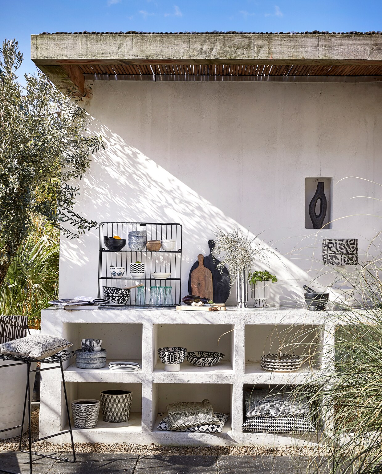 Metalen wandrek, 60x60 cm € 44,95, kussen op kruk € 22,50 (mrsbloom.nl). Metalen barkruk Wire, 47x42x82 cm(bxdxh) € 219,- (hkliving.com). In het rek: zwarte kommen Bold &amp; Basic, per 4 € 25,95, kommen Gradient, per 4 € 34,-, witte kommen Kyoto € 6,-(hkliving.nl), houten kommen € 12,95 (householdhardware.nl), witte gebloemde kom € 1,99, bloempot met golven € 3,99 (ikea.nl), schaal, per 4 € 33,56 (hkliving.com), Marokkaanse kommen Fex, per stuk € 55,50, houten lepel € 5,-, glazen Diabolo uit Marokko € 6,- (householdhardware.nl). Op het keukenblok: walnoothouten plank € 37,50 (householdhardware.nl), zwarte houten serveerplank € 42,95, leistenen snijplank € 24,95, gestreepte vaas Toppu van Oyoy € 69,95, pot op voet € 19,95 (livvlifestyle.nl). Spekstenen vijzel € 34,95 (dille-kamille.nl). Lampenkap Adir € 69,90 (ateliersem.nl). Organische vaas in nis € 51,95 (ptmd.nl). In de vakken: diepe borden Gradient, per stuk € 35,-, kommen met palmboom € 7,95, ontbijtbord met palmboom € 9,95 (hkliving.com), saladeschaal Fez uit Marokko € 129,-, grote bowl Fez € 55,-, borden Fez Assiette € 79,95 (householdhardware.nl), bloempot met ikatprint € 28,95, pot met geometrische print € 51,95, stoelkussen onder met grafische print € 45,95 (ptmd.nl). Tafellinnen € 64,95 (mrsbloom.nl). Kussen boven € 45,95 (ptmd.nl), kussen Stockholm € 19,99 (ikea). vtwonen 06-2023 | styling Kim Timmerman | fotografie Peggy Janssen