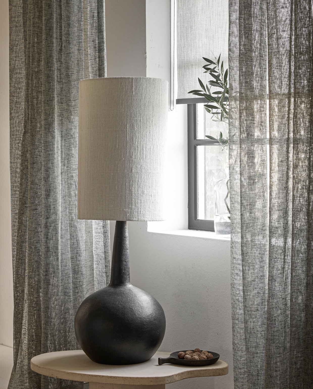 Inbetween gordijn op maat met enkele plooi in stof Weave (2712) kleur Brush, vanaf vanaf € 32,99 per strekkende meter. Lichtdoorlatend rolgordijn op maat in kleur antraciet (18015), vanaf € 128,19. M.m.v.: rawmaterials.eu (tafellamp Loulou XL), bogard.eu (salontafel Palet 80x50 cm in kleur Stone, afgewerkt in beton op MDF), vtwonen.be/shop (schaal met handvat van zwart mangohout). vtwonen 06-2023 | productie Tanja Saarberg | styling Marit Saladini Platjouw | fotografie Peggy Janssen
