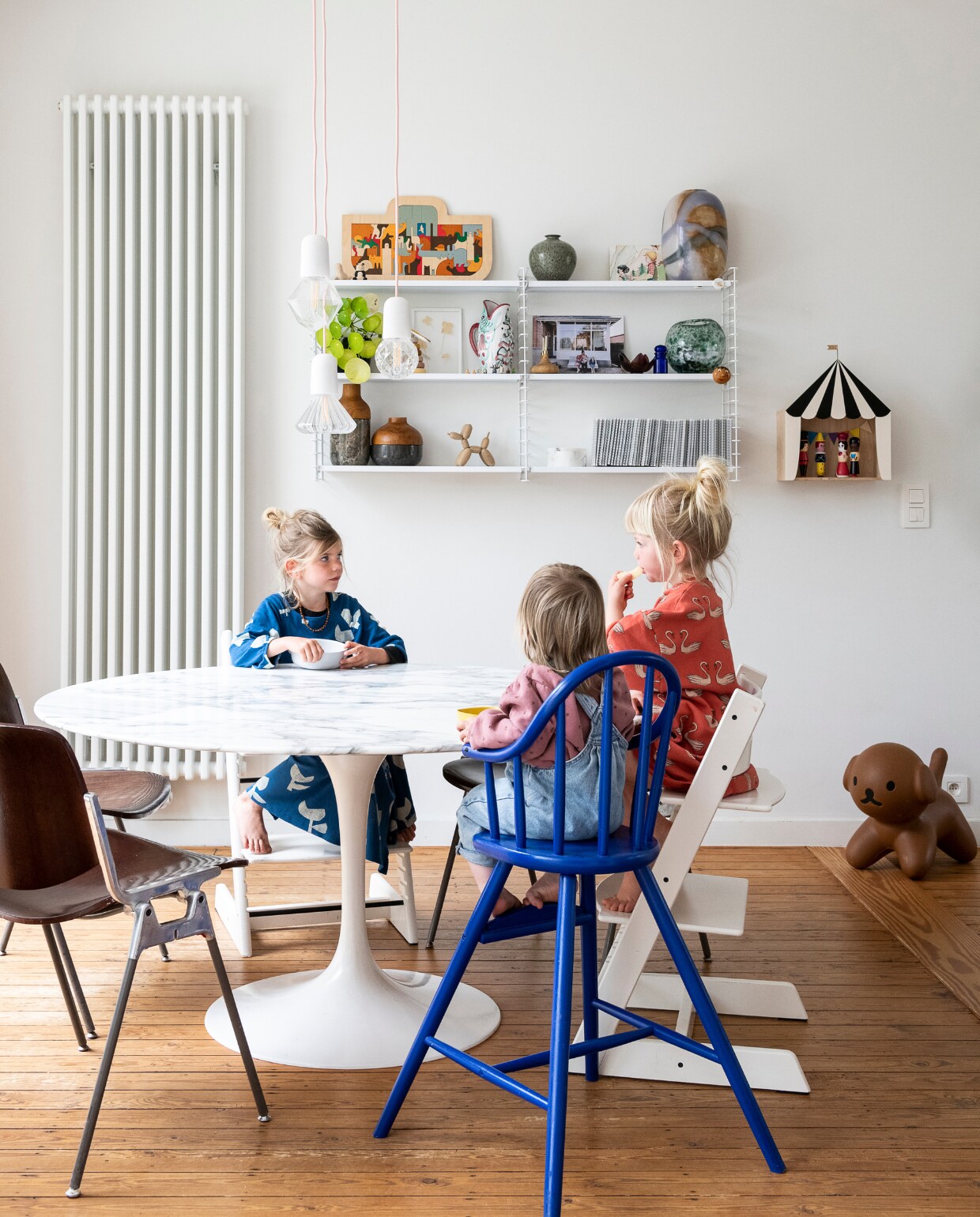 Marmeren eettafel Tulip van Eero Saarinen voor Knoll, witte meegroeistoelen Tripp Trapp, blauwe kinderstoel Ikea (zelf geverfd), bruine stoelen Castelli, lampen Zangra, wandrekken Tomado. Op wandrekken: kaarsenhouder A wilder home, vazen en potten Zara Home en H&amp;M, vis-waterkan Anna+Nina, houten puzzel Ark van Noach George Luck van Hape, ballonhond Boyhood, wanddecoratie circus UpWarsaw met daarin houten magnetische figuren van Vilac, zitje Snuffie Mr Maria. Verwarming Radson Delta. Vloer (originele) grenen planken met kleurloze olie. vtwonen 06-2023 | styling &amp; fotografie Jonah Samyn