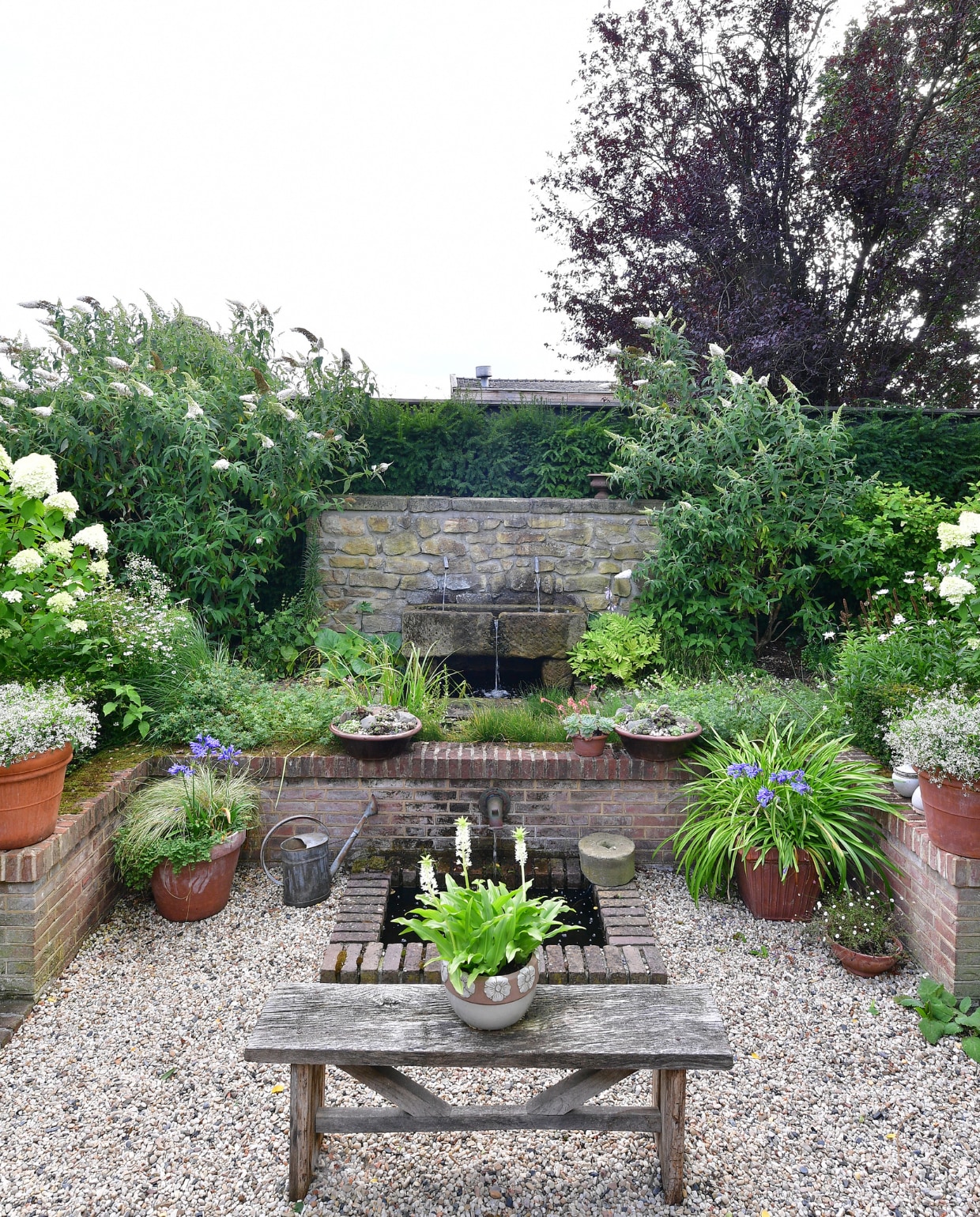 De zitkuil heeft links en rechts een trappetje. De pluimhortensia’s op de vier hoeken geven de kuil meer diepte, net als de euphorbia’s in de terracottapotten. Om dit gedeelte van de tuin het effect van een besloten stadstuin te geven, is gekozen voor een vooral wit met groene beplanting. Uit het muurtje stroomt water uit twee zinken pijpjes, ervoor is een bak gemaakt van Bentheimer zandsteen waar het water in klettert en overloopt in een vijvertje in de straat. vtwonen special tuin 04 | fotografie Sietske de Vries | styling Ina ter Haar