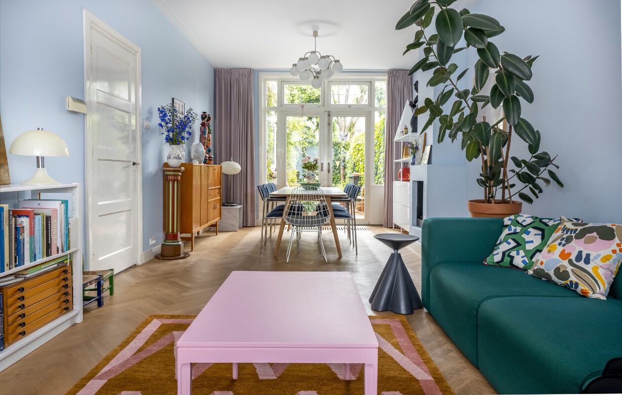 Muurverf S2.08.79 van Sikkens, houten visgraatvloer was al aanwezig, gordijnen Hema, hanglamp Boulanger, stellingkasten Type 663 Gispen. Dressoir, ladeblok, stalamp en beschilderde rieten stoelen vintage. Vloerkleed Folding Ribbon van Nordic Knots, oude pilaar uit een kerk, marmeren bijzettafel de Witte Eenhoorn. Tafel Bella van Hay, blauwe stoelen Gijs van der Sluis, draadstoelen Wire van Pastoe. Kan op pilaar en vaas op tafel souvenirs. Koolbladschaal Bordallo Pinheiro, bijzettafel Philippe Starck, salontafel Decennia Design, 3-zitsbank Mags van Hay, kussens Marimekko en Hay. Plant is een Ficus. vtwonen 07-2023 | fotografie Henny van Belkom