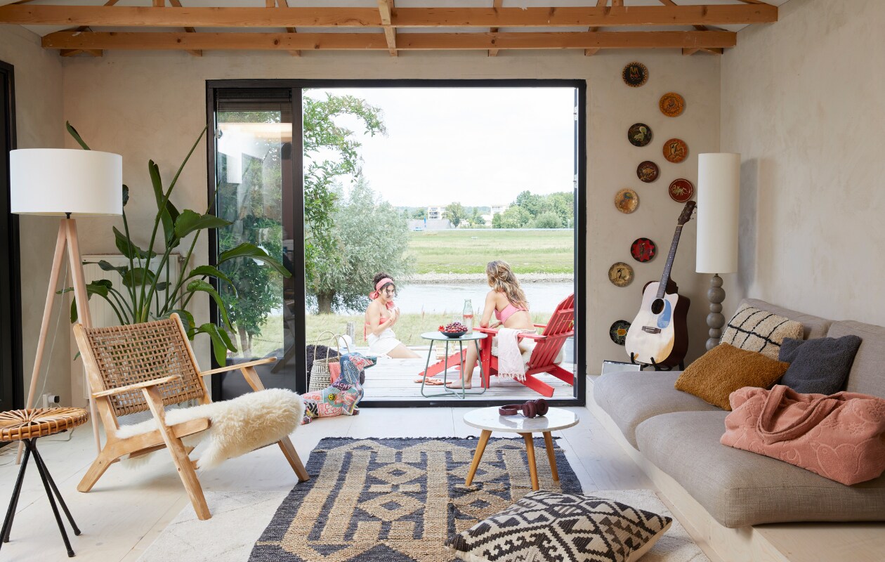 Fauteuil Grignoon van Kave Home, lampen gemaakt voor OT Design, rieten bijzettafel erfstuk. Kunstwerk teckel gemaakt door Lisa. Vloerkussen gemaakt van stof van Chivasso. Handgeschilderde wandschotels meegenomen uit Sumatra, grijze kussens oude bank. Mand uit Afrika, salontafel Zuiver, kussens en tafel buiten Karwei, strandtas meegenomen van Ibiza, rode Adirondack-stoel vidaXL. Underlayment vloer gelakt. Overig onbekend. vtwonen 08-2023 | styling Linda van der Ham | fotografie Simone de Geus