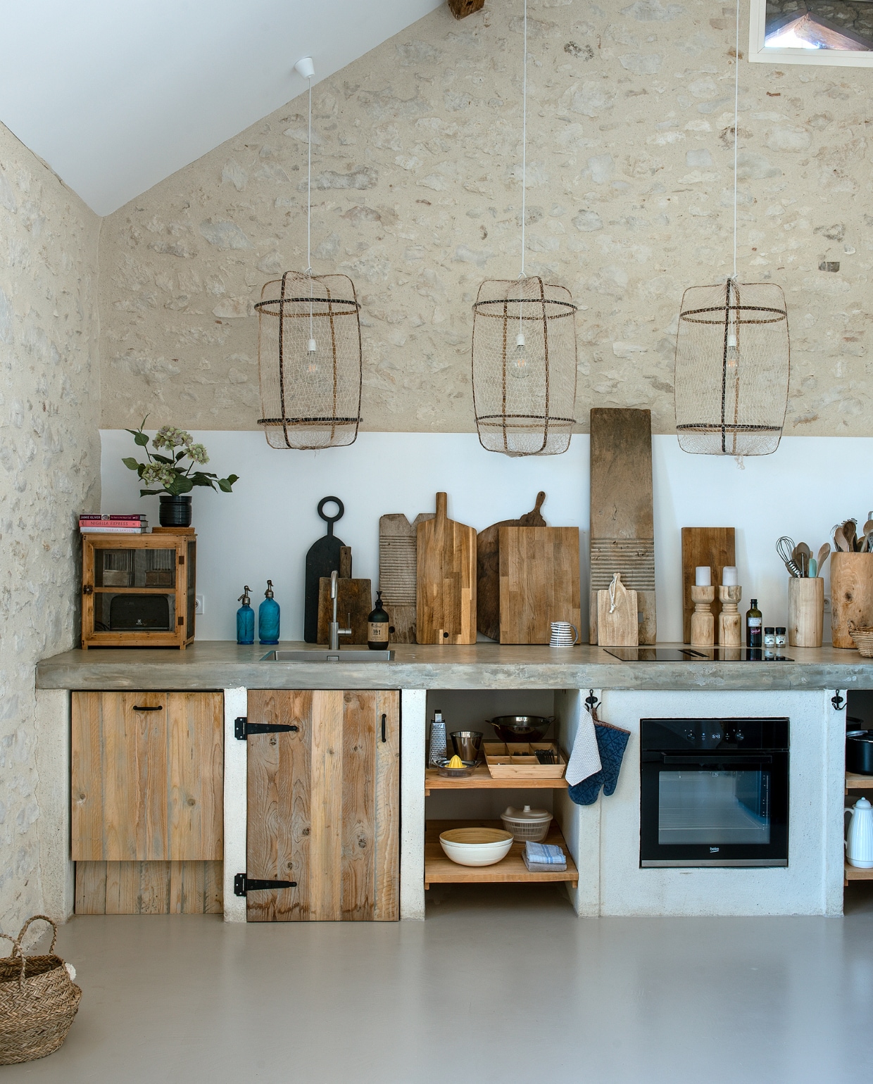 Keuken eigen ontwerp met betonnen blad en deuren van oud hout. Hanglampen @just_bysan. Houten kandelaars Bloomingville. Houten potten en planken, vliegenkastje en spuitflessen van verschillende brocantezaken. Mand en keukenspullen grotendeels Ikea. Overig onbekend. vtwonen special binnenkijken 06-2023 | fotografie Ton Bouwer | productie Monique van der Pauw