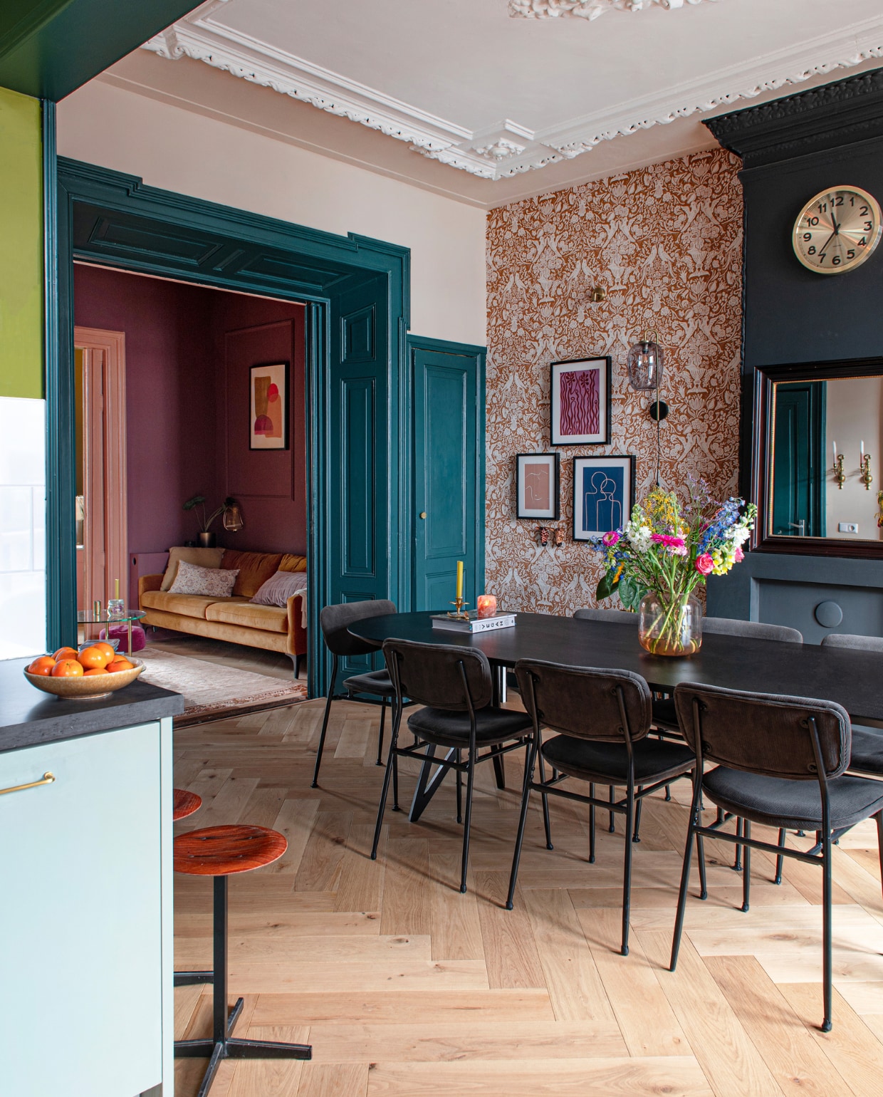 Behang Karwei, klok Karlsson, eettafel Studio Henk, stoelen Wonenmetlef, wandlamp Marktplaats. Muurverf schouw Off-Black van Farrow &amp; Ball, verf wand en plafond IG KH M-058 van Karwei. Visgraatparket Blank Geolied Eiken van vtwonen via Karwei Paarse print Etsy, blauwe print Atelier Andrea, roze print Bo Sterenberg. Kandelaar À La Collection, fruitschaal Sissy-Boy, barkrukken en salontafel vintage, bank Sofacompany, kunstwerk Enkel Art Studio, vloerkleed BePureHome, gebloemd kussen Karwei, geel kussen Essenza. Overig onbekend. vtwonen 09-2023 | productie De Binnenkijkers | styling Jeske Weel | fotografie Caroline Coehorst