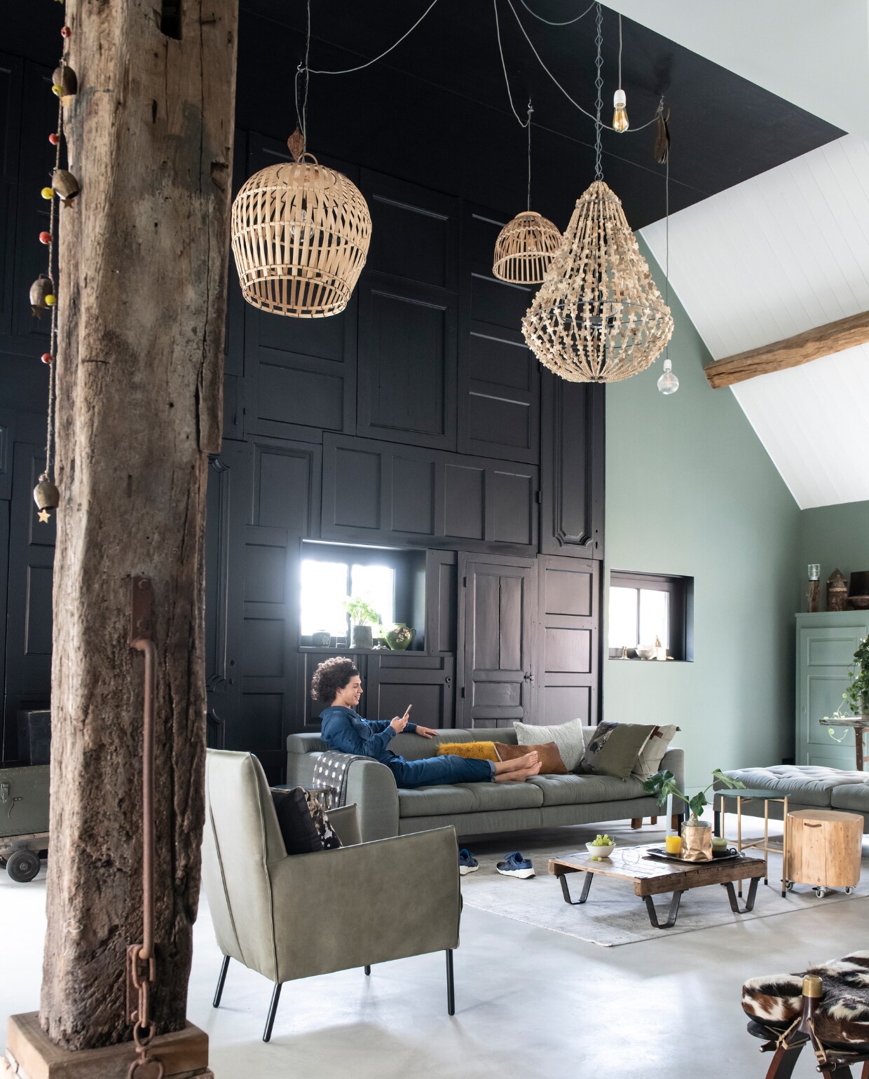 De hoogte van de voormalige boerderij verleidde Cindy en Alex tot een radicale ingreep. Zwart geverfde paneeldeuren (After Dark) creëren een markante wand in combinatie met de kleuren Leaf en Ash van Stapelgoed. Bank vtwonen (met eigen stof bekleed), stoel Stapelgoed (niet meer te koop). De twee bamboe lampen en de Cross lamp (Stapelgoed, niet meer te koop) zijn bevestigd aan oude katrollen. Vloer gietbeton, de lage pallettafel maakte Alex zelf. vtwonen 11-2019 | styling Esther Loonstijn | fotografie Louis Lemaire