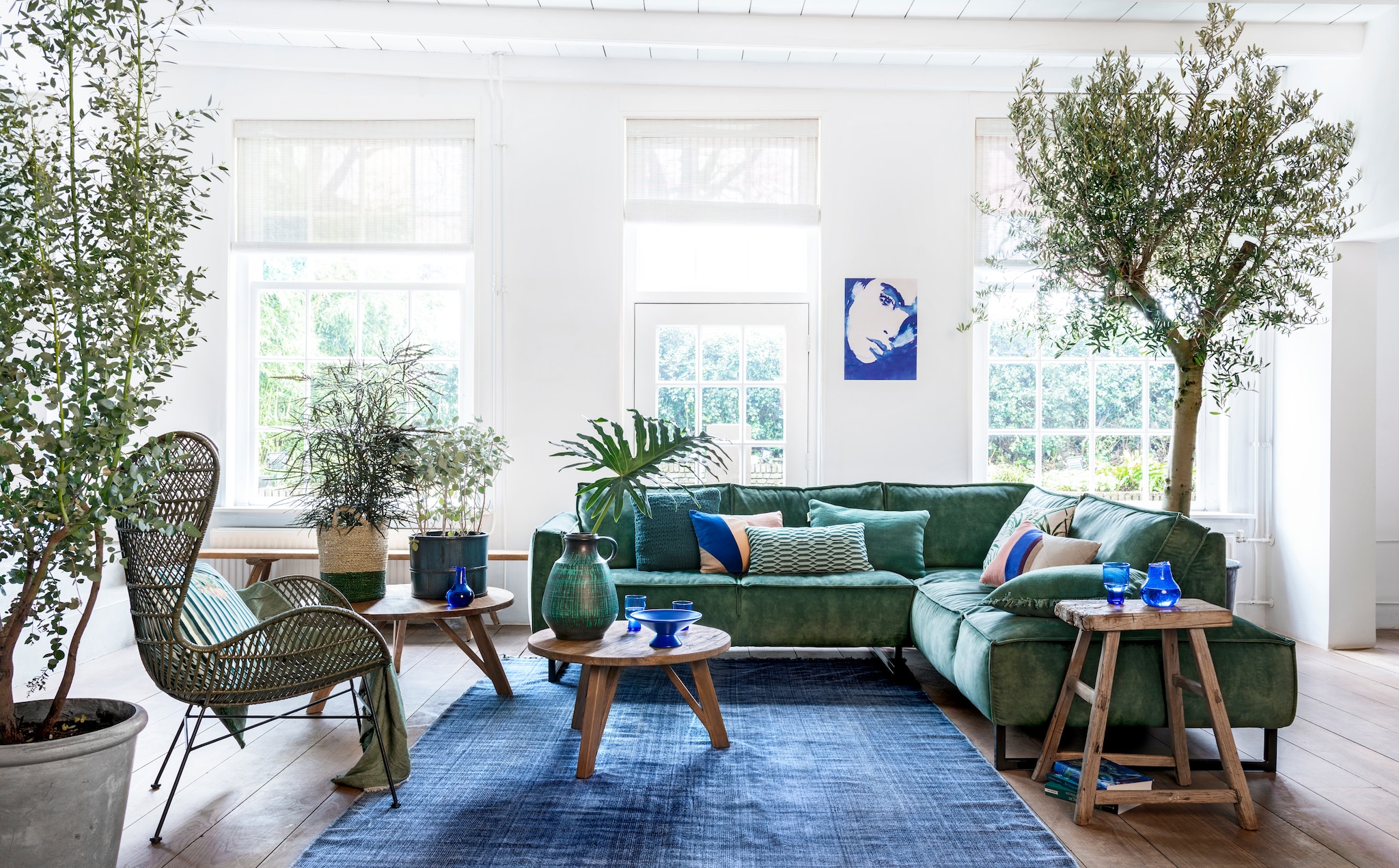 Hoekbank Kick € 1650,-, grote salontafel € 449,-, kleine salontafel € 299,-, rotan fauteuil Egg Chair € 395,- (hetkabinet.nl). Gestreept kussen op fauteuil € 29,99 (sissy-boy.com). Groene linnenmix plaid € 39,99 (hm.com). Vloerkleed Vietnam Days, Stonewashed Blue Cotton Block Print, 200x300 cm € 398,- (aaimadewithlove.nl). Boompjes: olijfboom en Eucalyptus (kwekerij). Naturel-groene mand € 59,95 (manggisliving.nl) met plant Schefflera Elegantissima (tuincentrum). Blauw houten gelakte plantenpot € 49,- (hetkabinet.nl) met plant Kalanchoë (tuincentrum). Blauwe glazen karaf per 3 € 26,95, blauwe fruitschaal van keramiek € 29,95, hoge blauwe glazen per stuk € 8,95, lage glazen per stuk € 6,95 (hkliving.nl). Grote vintage vaas € 65,- (mippies.com). Kussens op de bank van links naar rechts; vlakken multicolour € 39,95 (hkliving.nl), langwerpig groen gedessineerd € 29,99 (sissy-boy.com), groen stonewashed kelim € 49,95 (hetkabinet.nl), met bladprint € 9,99 (hm.com), met vlakken Patch € 39,95 (hkliving.nl), groen stonewashed Kelim € 35,- (hetkabinet.nl). Poster € 29,95 (hkliving.nl). Bankje, grote plantenpot links, houten kruk en boeken privébezit. vtwonen 05-2019 | Styling Moniek Visser | Fotografie Stan Koolen
