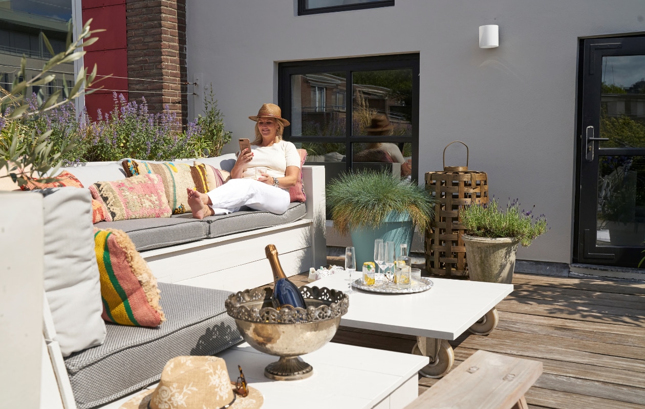 Loungemeubel en tafel op wielen eigen ontwerp. Kruk en geweven lantaarn Loods 5. Koperen buitendouche Seletti. Kussens op de bank Tim’s Kussens, kleurige kussens Bohemian du Maroc. Zilveren schaal op voet ooit bij de Bijenkorf gekocht. vtwonen 08-2020 | productie Features &amp; more | styling Wilma Custers | fotografie Luuk Geertsen