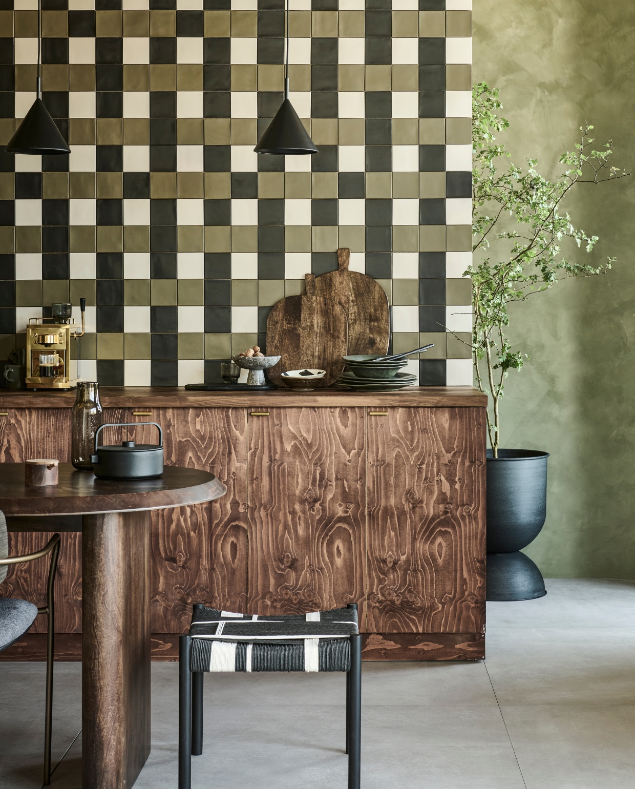 Op de wand mix van tegels Chop Kaki, Chop Nero en Chop Bone, 10x10 cm, per m²  € 79,90. Op de vloer tegels Raw Grey Mat, 120x120 cm, per m² € 92,50.M.m.v. pure-original.com (muurverf Olive Drab), basiclabel.nl (mangohouten tafel Ellips van vtwonen, kruk Jessy van Woood), hm.com (mangohouten snijplanken, marmeren etagère, zwarte snijplank, karaf, mangohouten lepels), debijenkorf.nl (schaaltjes van HKliving), zarahome.com (borden, houten suikerpot, mokken), vtwonen.nl/shop (bloempot van Ferm Living, stoel van Kave Home, hanglampen van HKliving, theepot van Serax), sjostrandcoffee.com (espressomachine). vtwonen 09-2023 | productie Tanja Saarberg | styling Sanne Ketelaar | fotografie Sjoerd Eickmans