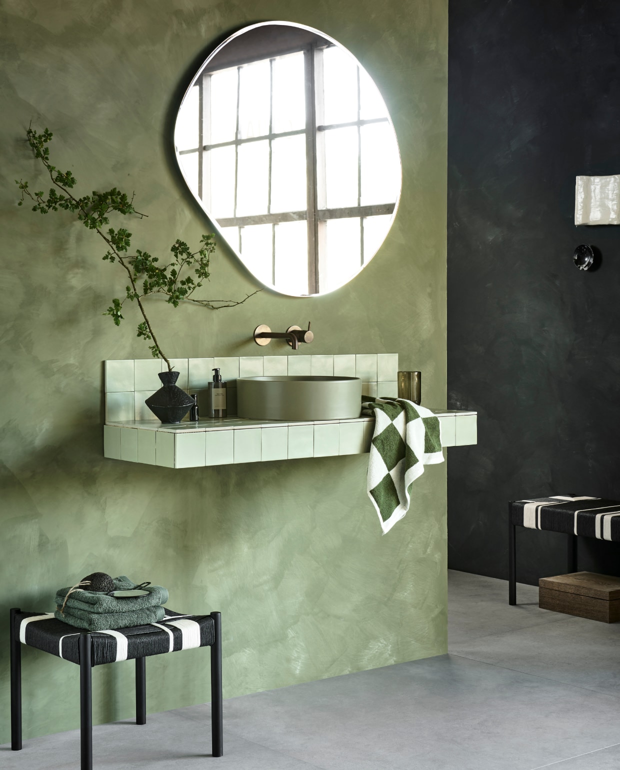 Op het wastafelmeubel tegels Chop Salvia, 10x10 cm, per m² € 79,90. Op de vloer tegels Raw Grey Mat, 120x120 cm, per m² € 92,50.M.m.v. pure-original.com (muurverf Olive Drab en Dark Forest), vtwonen.nl/shop (spiegel Pond van Ferm Living), tegeldepot.nl (waskom van Salenzi, wastafelkraan van Hotbath in geborsteld koper), basiclabel.nl (kruk Jessy van Woood, bank Jessy van Woood), mooswillemsen.com (kunstwerk), zarahome.com (vaas, handcrème, groene handdoeken), hm.com (glas, handspiegel), debijenkorf.nl (geblokte handdoek van Hay). Overig privébezit. vtwonen 09-2023 | productie Tanja Saarberg | styling Sanne Ketelaar | fotografie Sjoerd Eickmans