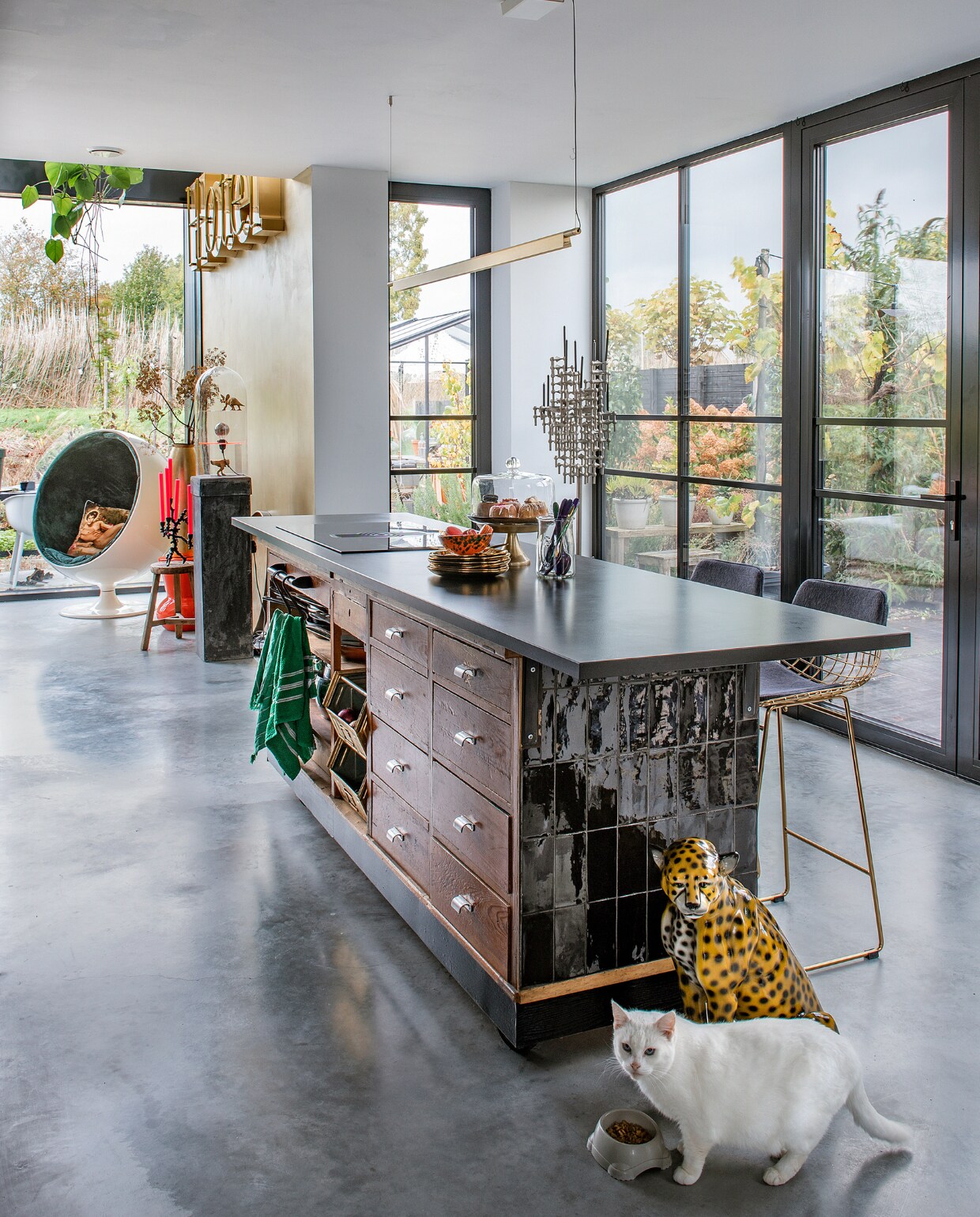 Toonbank Werkplaats 35, granieten werkblad Keukenloods, tegels Tegelsonline. Gietvloer Willem Designvloeren. Gouden borden en oranje kruk De Kaardebol. Bestek Sluiz, barkrukken HKliving, letters hotel Eigenaar 8, houten kruk Nijhof. Fauteuil Ball Chair van Eero Aarnio, kandelaar Stoff Nagel, luipaard vintage. Overig onbekend. vtwonen 10-2023 | fotografie Ton Bouwer