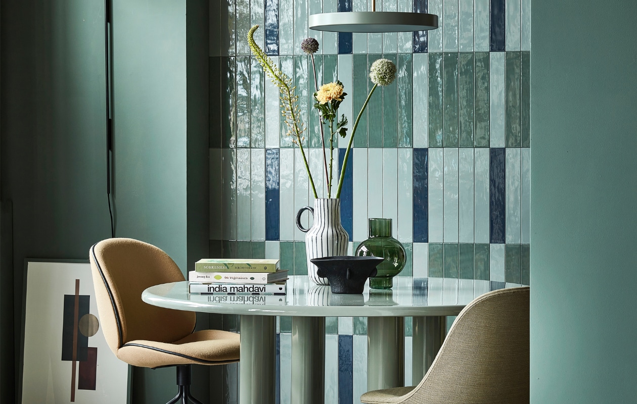 Glanzende wandtegels Marrakesh van vtwonen in kleuren Ocean Blue, Sea Green en Army Green, 6,5x40 cm, per m2 € 70,-, stalen wandlamp Arum van Ferm Living € 397,-, aluminium hanglamp Asteria van Umage € 205,95, groene glazen vaas Tummy van Urban Nature Culture € 25,95 (vtwonen.be).Muurverf in Green Smoke No.47, per 2,5 liter € 97,- (farrow-ball.com). Eettafel Smalto van geëmailleerd staal van Knoll, 137x73 cm (Øxh) € 4888,50 (justhaasnoot.nl). Stoel links Beetle van Gubi in Divina 3, staal en textiel € 1249,-, stoel rechts Harbour Side Dining chair van Menu, kunststof en staal € 660,- (hellochair.nl). Zwart-witte kan € 24,99, zwarte schaal € 24,99 (hm.com). Poster Shapes of Colour 03 van Paper Collective, 50x70 cm € 49,-, eikenhouten lijst Quadrum, 50x70 cm € 64,95 (loods5.nl), meegeschilderd met de muur. vtwonen 10-2022 | styling Kim van Rossenberg | fotografie Peggy Janssen