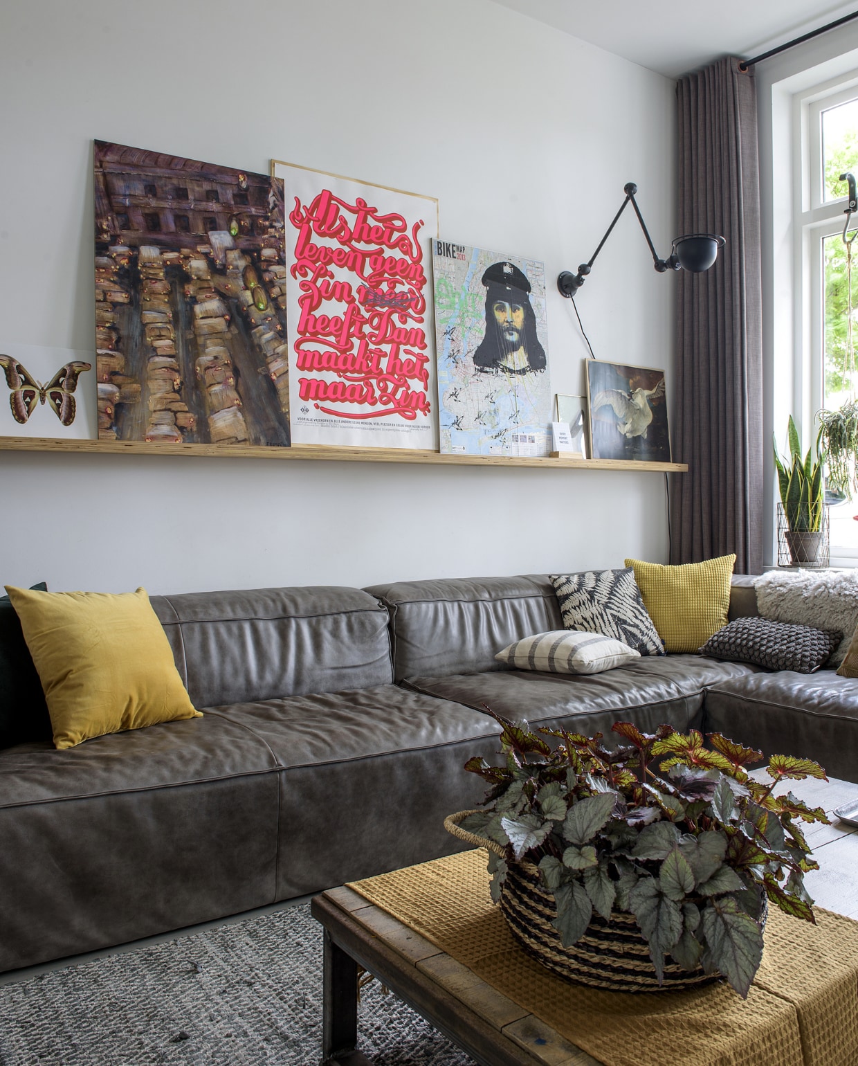 De modulebank van Het Anker loopt helemaal door voor het raam en biedt zo ruimte aan het hele gezin, plus zoete inval. De kussens zijn van Ikea (geel), HK Living (bladmotief), Hema (streep), het groene kussen en vloerkleed zijn van Groene Vingers Delft. Lamp van Jieldé. De koffietafel is gemaakt van een steenschot en een onderstel gelast door een familielid. Op de wandplank staan een poster met roze tekst van Studio Boot, een fietskaart van New York met Jezus-afbeelding meegenomen uit New York, een schilderij van Wendy’s broer Frank en een vlinderfoto van Liljebergs, gekocht in Kopenhagen. vtwonen 01-2019 | fotograaf Ton Bouwer | styling Wendy Romeijn en Monique van der Pauw
