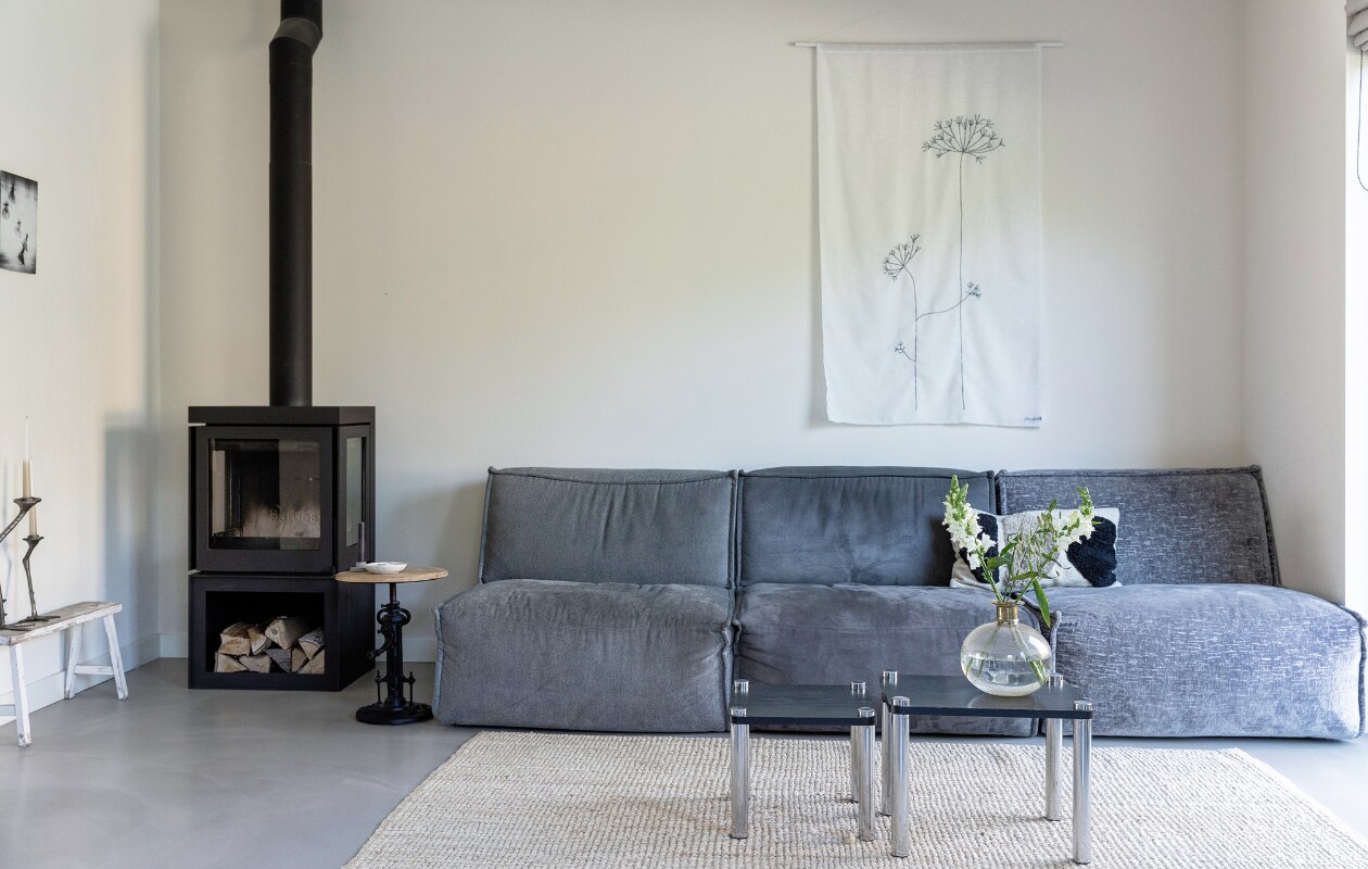 Elementenbank Evo van De Troubadour, linnen wanddoek Huize Jolie, vloerkleed Marktplaats, bijzettafel vintage. Kaarshouder en theelichthouder Twan van Leeff. Kussen H&amp;M Home, vintage bijzettafels Van Dijk &amp; Ko, vaas Artemis van Gifts Amsterdam, houtkachel Box Panorama 65 van Barbas Bellfires, houten bank Kringloopwinkel, kaarsenstandaards Voet van PaperBag, print Dutch Angels van Julien Oncete van Kunstkopie. vtwonen 09-2023 | styling Ilona Jongepier | fotografie Dana van Leeuwen