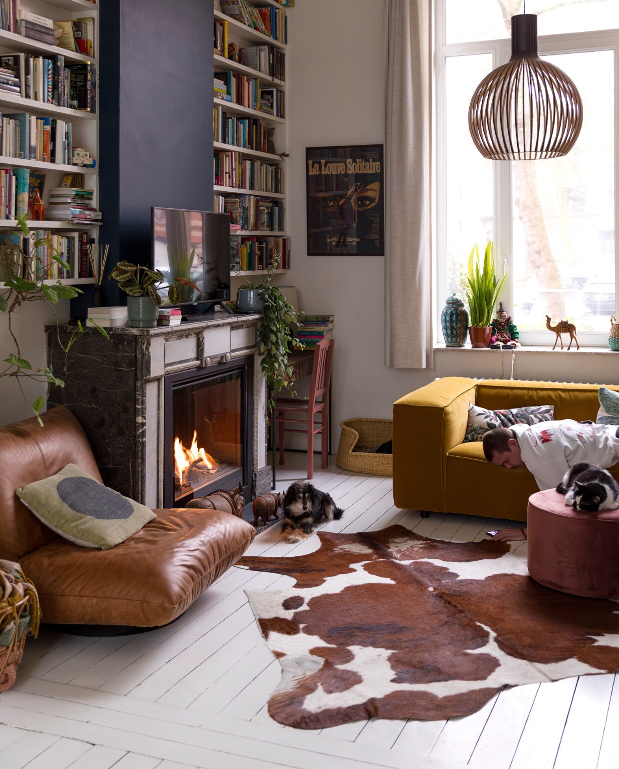 Muurverf schouw Hague Blue van Farrow &amp; Ball, verf overige wanden en vloer Ral 9010. Gordijnen en planken op maat laten maken.  Scheepsfauteuil Woontheater, haard 4M-Fire, potten en planten Van Uytsel. Hanglamp Octo van Secto, potten in vensterbank rommelmarkt, stoel kringloop, vloerkleed koevel van de markt. Bank Dunbar van Fést, poef Sara van Woood, hondenmand Dille &amp; Kamille, poster brocante. Kussens Sissy-Boy en HKliving. Overig onbekend. vtwonen 10-2023 | styling Ilona Jongepier | fotografie Dana van Leeuwen