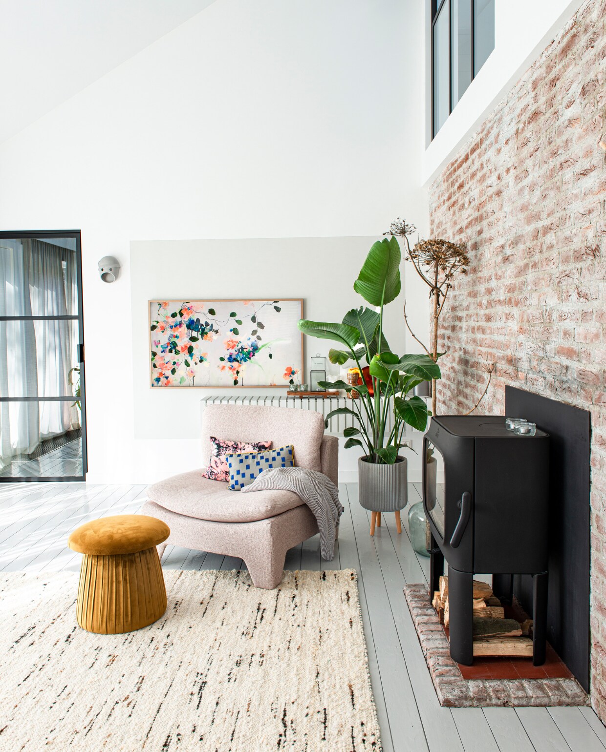 Stalen raam en deur Steellife. The Party Wall Lamp Coco van Moooi, plant met pot Intratuin. Televisie The Frame van Samsung, kader op de muur erachter kalkverf kleur parelwit van Kalk&amp;Co. Wandkast van latten zelfgemaakt en geschilderd met dezelfde kalkverf als het wandvlak. Fauteuil, kussens en vloerkleed  HKliving. Poef Mushroom van Sissy-Boy. Houtkachel Jøtul van Nijhof. vtwonen special DIY 6-2022 | productie De Binnenkijkers | styling Jeske Weel | fotografie Caroline Coehorst