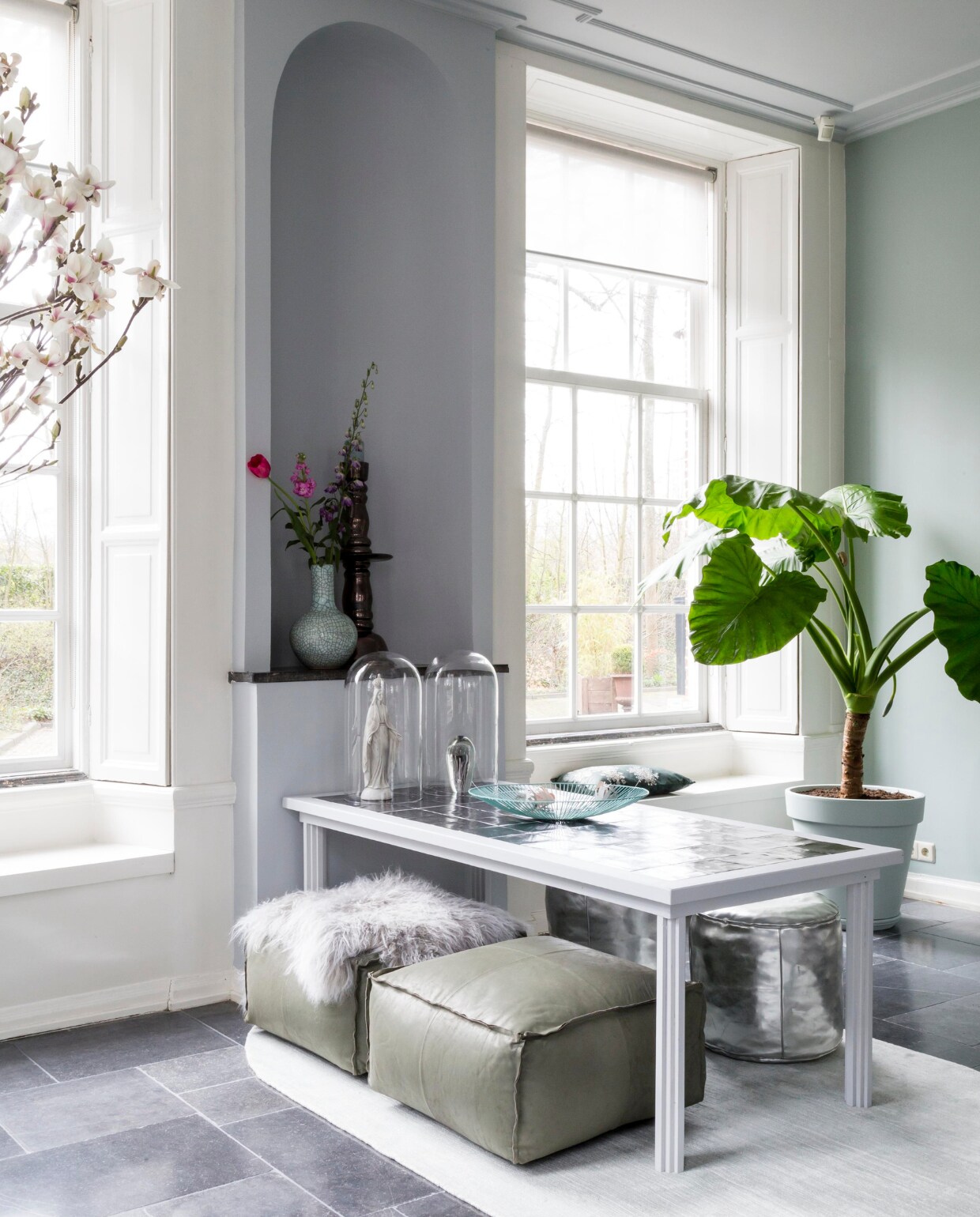 Grijze muurverf Wild Dove, groene muurverf Early Dew (flexa.nl). Grote plant (greenlifestylestore.nl) in zelfgeschilderde terracotta pot (tuincentrum). Tafel (com-anders.nl). Ovalen vloerkleed in zachtgroen van HK Living, 150x240 cm, tegels in tafelblad Villa Platinum Metallic van vtwonen, lichtblauwe schaal van Serax (vtwonen.nl). Grijsgroene vierkante leren poefs, glazen stolp (casavivante.com). Ronde zilverkleurige poefs, kandelaar (householdhardware.nl). Mariabeeld (clayre-eef.nl). Glazen presse-papier met kwal (hkliving.nl). Craquelé vaas Mint (polspotten.nl). Overig privébezit. Weer verliefd op je huis seizoen 10 aflevering 8 | styling Marianne Luning | fotografie Barbara Kieboom