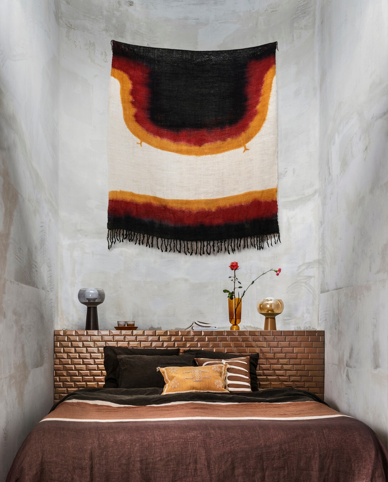 Wollen, met de hand geverfd wandkleed (householdhardware.nl). Linnen dekbedovertrek van Chivasso (jab.de). Bruin-wit kussen, kussen met borduursel (zenza.nl). Op bed een kanoko, ofwel een linnen tie-dye doek van Osborne &amp; Little (wva.nl). Lampen Mushroom van BePureHome, vaas van Hay, mokken van HKliving (vtwonen.be). vtwonen 02-2022 | styling Marianne Luning | fotografie Stan Koolen