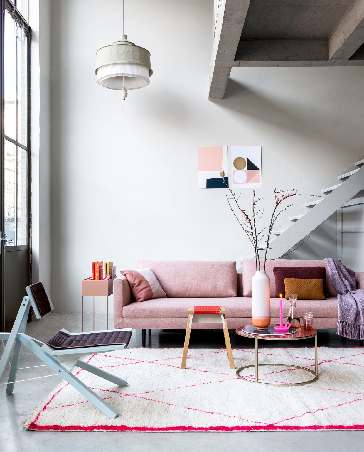 Muurverf RAL5800 (Sikkens). Bank Etcetera, gestoffeerd in roze stof Synergy van Eyye, 238x66x89 cm (lxhxd) (Design on Stock). Houten krukje met gewatteerde zitting, fauteuil, tentlamp met porseleinen kraal (Scheublin &amp; Lindeman). Plantenbak (met boeken erin) Ferm Living, okergele en bordeauxrode kussens op de bank (Kolk en Kolk). Rode portemonnee van Freitag, karaf, glazen, boeken (Piet Hein Eek). Messing bijzettafel met donkerroze keramisch blad (Pols Potten). Berber karpet met fluoroze strepen, 200x300 cm (Household Hardware). Plaid (Malagoon). Two tone roze kussen, kaars in kandelaarvorm (Ontwerpduo). Dipvaas Nude, platte schaal van porselein (Studio Harm en Elke). Posters aan de muur (Tom Pigeon). vtwonen 06-2018 | Styling Marianne Luning | Fotografie Stan Koolen