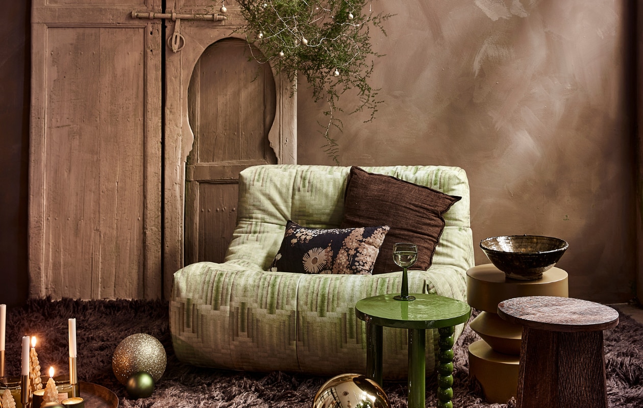 Fauteuil Lazy Lounge Chair, 105x105x75 cm (bxdxh), bruin hoogpolig vloerkleed Fluffy in Espresso, wol en katoen, 200x300 cm (hkliving.nl). Groene acaciahouten bijzettafel Hava, okergele metalen bijzettafel Bora, mangohouten bijzettafel Lux, keramieken theelicht, kerstboomkaarsjes (loods5.nl). Gouden kunststof kerstballen (kerstversiering.nl). Keramieken schaal op tafel (householdhardware.nl). Goudkleurige kandelaars laag, hoog (hema.nl). Groene glazen kerstbal, per 6 (bol.com). Gouden glazen kerstballen (xenos.nl). Krans zelfgemaakt. Overig privébezit. vtwonen 13-2023 | styling Moniek Visser | fotografie Peggy Janssen