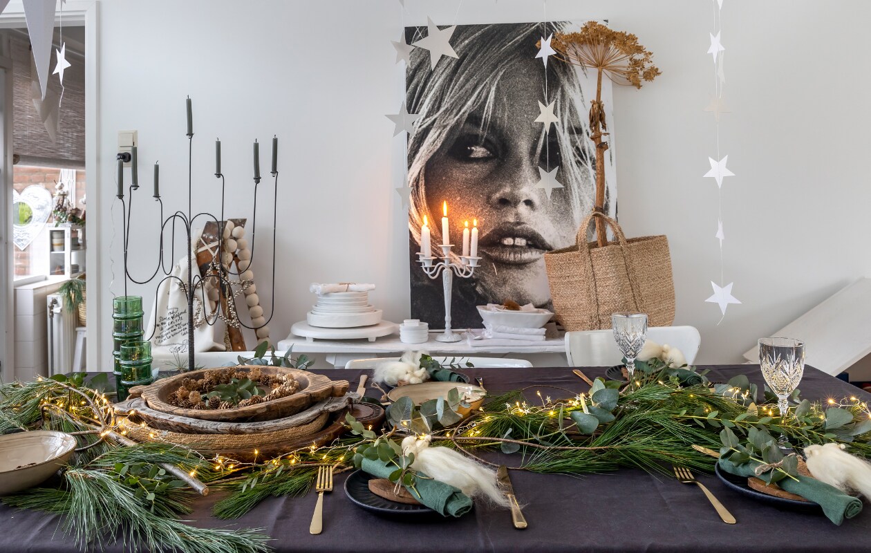IJzeren zevenarmige kandelaar De Tuinkamer brocante. Mand H&amp;M. Witte ijzeren kandelaar, eetkamerstoelen, houten schilderstrap Marktplaats. Grijs tafelkleed H&amp;M. Gouden bestek Albert Heijn en Sissy-Boy. Witte houten schalen Action (zelf geverfd). Tas Sukha. Groene takken markt. Houten schalen Marktplaats en kringloop. Groene glazen Household Hardware. Groene servetten Søstrene Grene. Zwarte borden Sissy-Boy. Beige schalen Xenos. Wijnglazen kringloop. Sterren Aliexpres. Engelen op borden zelfgemaakt. Overig onbekend. vtwonen 13-2023 | fotografie Henny van Belkom | tekst Ina Babbé