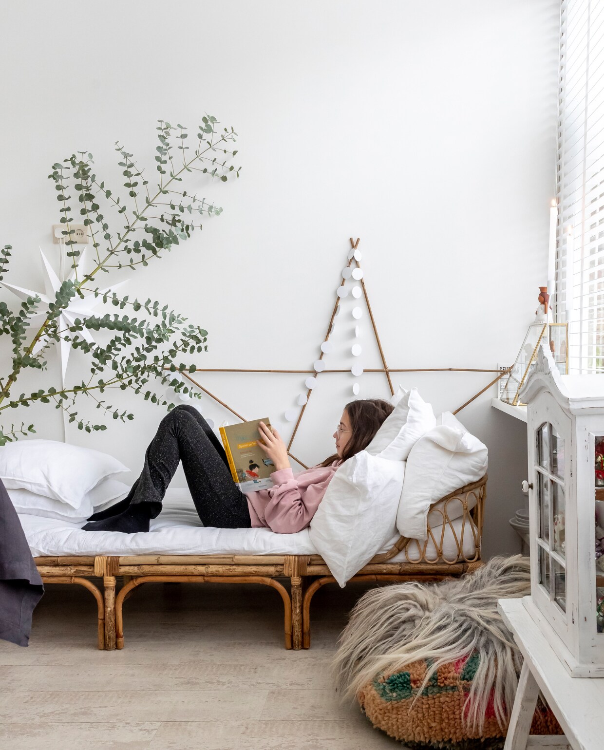 Olivia op een daybed van Karwei. Kussens daybed H&amp;M. Vitrinekast Marktplaats. Poef kringloop. Schapenvacht Vacht van Vilt. Witte sterren en kerstslingers Aliexpress. Bamboe ster zelfgemaakt. Eucalyptustak markt. Houten bank Marktplaats. Gouden vitrine op vensterbank Sissy-Boy. vtwonen 13-2023 | fotografie Henny van Belkom | tekst Ina Babbé