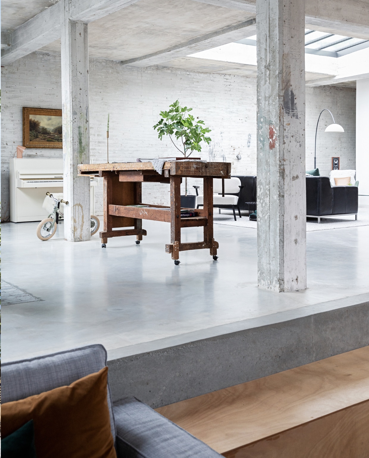 Vloer gevlinderd beton. Hardstenen waaltjes Spotted Bluestone. Werkbank erfstuk meubelfabriek. Kinderfiets Trybike. Witte fauteuil Ode Lounge Chair van Studio Henk. Bank Ikea. Booglamp Venled. Piano en schilderij erfstukken. Lamp op piano Elleke Rutten. vtwonen 01-2022 | styling Ilona Jongepier | fotografie Dana van Leeuwen