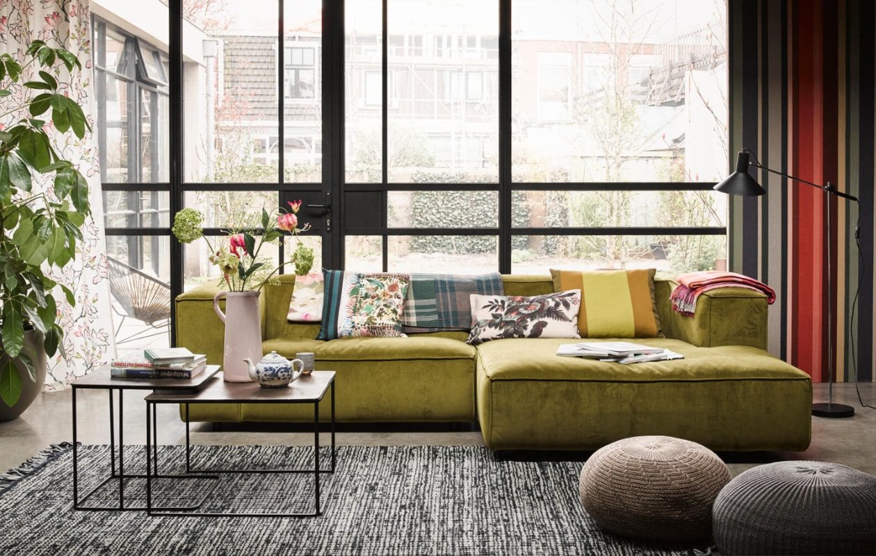 Gestreept behang Masterpiece nr. 358021 (Eijffinger). Bank Dunbar (Loods 5). Vloerkleed, poef (Ikea). Messing bijzettafels (HK Living). Gebloemd gordijn Kimono Blossom van Designers Guild (Wilhelmine van Aerssen). Op de bank v.l.n.r. plaid met bloemdessin Delft Flower Tuberose, kussen Herbier Jonc van Christian Lacroix (Wilhelmine van Aerssen), geruite plaid (Pols Potten), gebloemd kussen (HK Living), kussen brede streep (Loods 5), geruite plaid Balintore Zinnia van Designers Guild (Wilhelmine van Aerssen). Staande lamp (Neef Louis). Op tafel: roze kan, kopje zwarte streep (Loods 5). Buiten: stoel Acapulco van Fair Furniture (vtwonen.nl). Theepot privébezit. vtwonen 07-2018 | Styling Fietje Bruijn | Fotografie Alexander van Berge