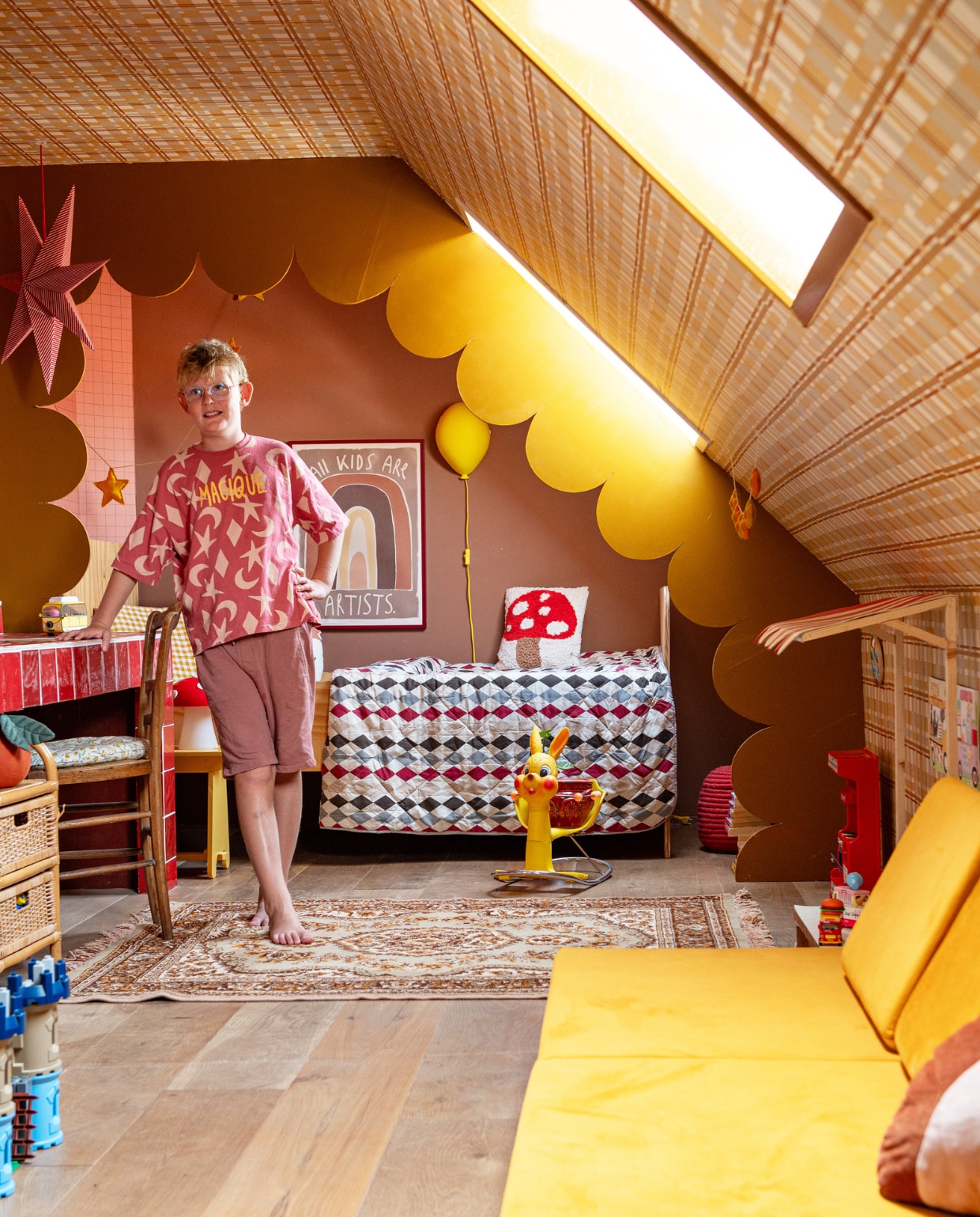 Muurkleur Affogato van Little Greene, behang Lilipinso, bedombouw zelfgemaakt en geschilderd in Bombolone van Little Greene. Bedsprei vintage, poster StudioLoco, ballonlamp Ikea, paddenstoelkussen Urban Outfitters, vloerkleed Hipstyling, wipstoel vintage via Yay Sugar Pie, houten marktkraam zelfgemaakt, gele bankkussens Funzy. Overig onbekend. vtwonen 01-2024 | styling Linda van der Ham | fotografie Caroline Coehorst