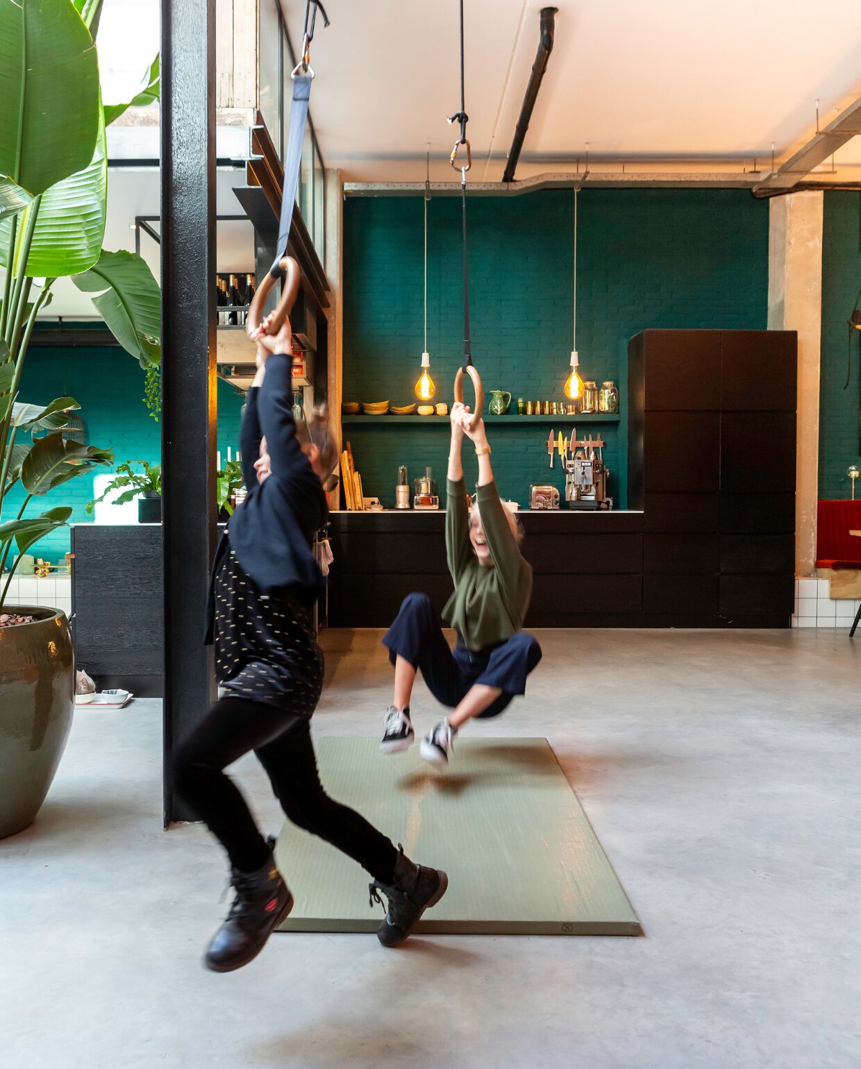 Lane en Kate slingeren aan de gymnastiekringen in hun loft-huis in Haarlem, USA. Keuken van Kvik met wit kunststof blad en zwarte frontjes met voelbare houtstructuur. Rek boven de bar gemaakt door Special Metal in Alkmaar. Theepot met koperen schild Bredemeijer, koffiemachine Buscaglione. Keramieken schaaltjes gemaakt door Marieke, Marokkaans glaswerk uit Marrakech. Hanglampen gemaakt van oude bakelieten fittingen met led-lampen. Ringen cadeau gekregen van een gymnastiekleraar. vtwonen 10-2020 | Fotografie Henny van Belkom