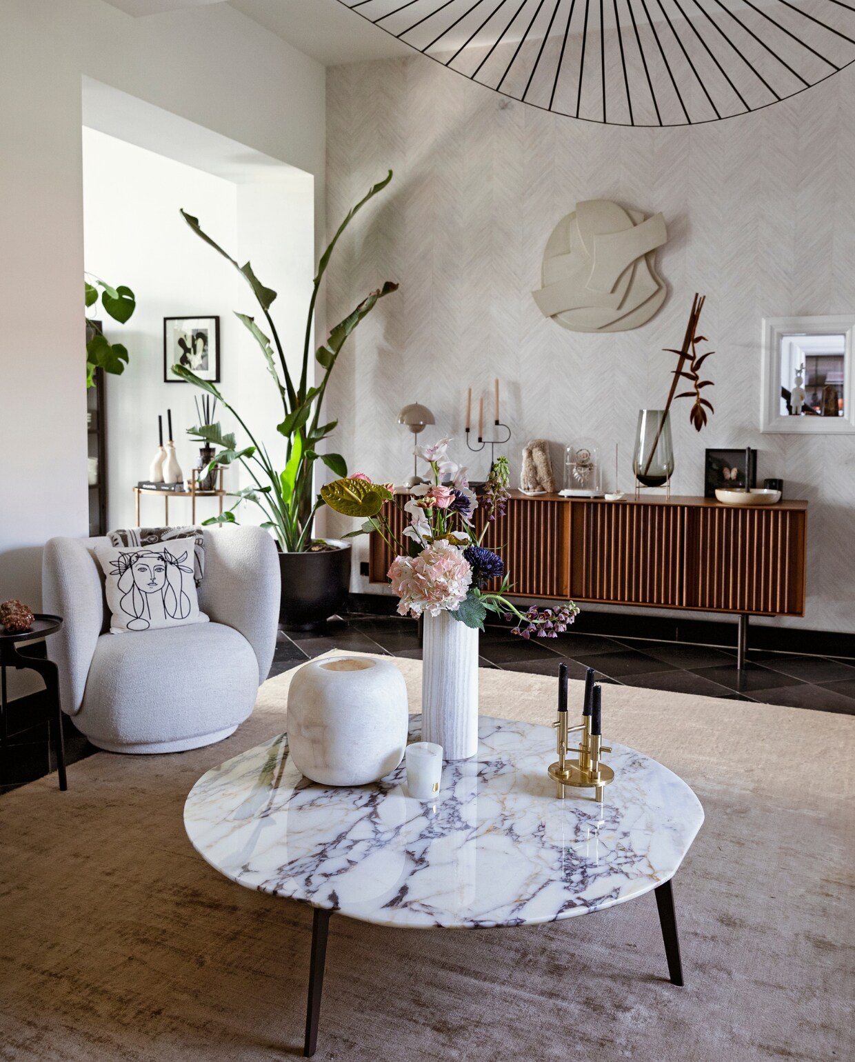 Plafondlamp Petit Friture via Interior Playground. Natuurstenen kerkvloer. Kleed Benuta. Marmeren tafel Papadatos via Decor Amsterdam. Witte vazen en kaars Albaster via Graanmarkt 13. Bloemen Van Esch Floral. Goudkleurige kandelaar en kandelaar rechts op kast Fritz Hansen via Loft 76. Bouclé stoel en kandelaar Ferm Living. Kussen Leclaireur. Bijzettafel Casa Gitane. Daarop: lamp &amp;Tradition. Kristal Earth Mineralen. Kunstwerk stolp Neeltje Geurtsen. Vaas Menu. Tak Joy Flowers. Aan de muur kunstwerk Atelier de Rijk. Trolley BePureHome. Daarop: kandelaars Anissa Kermiche. Geurstokjes Baobab. Schilderij Cate Adriana. Plantenpot &amp;Tradition. Behang Selva van Arte. Muurverf wit ral 9010. vtwonen 11-2021 | Styling Ilona Jongepier | Fotografie Dana van Leeuwen | Visagie Djolien de Kreij