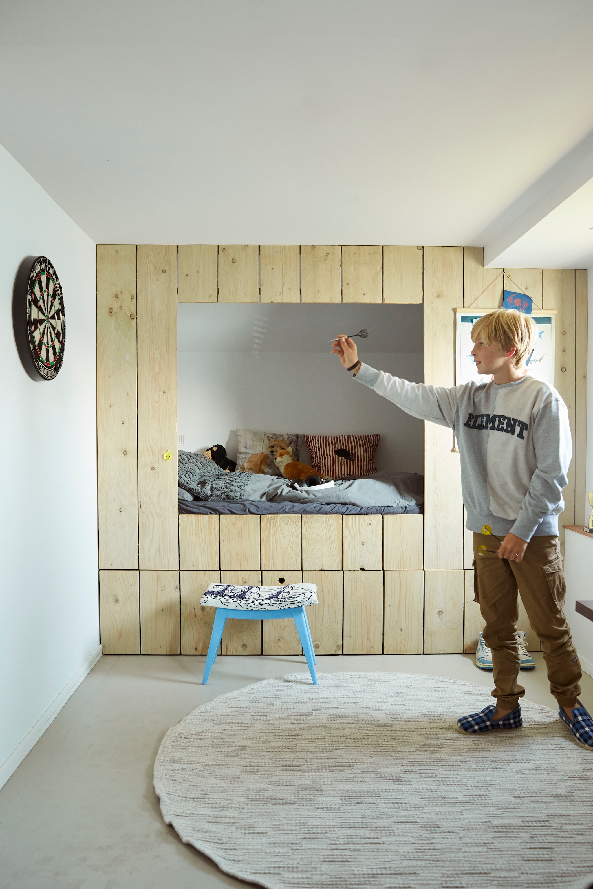 Dartbord tweedehands, zandcement vloer met betonverf, steigerhouten bedstee zelfgemaakt, onderlaken Hema, kussens Anna + Nina, knuffel Blokker. Overig onbekend. vtwonen 03-2024 BK Monnickendam | styling Marianne Luning | fotografie Tjitske van Leeuwen