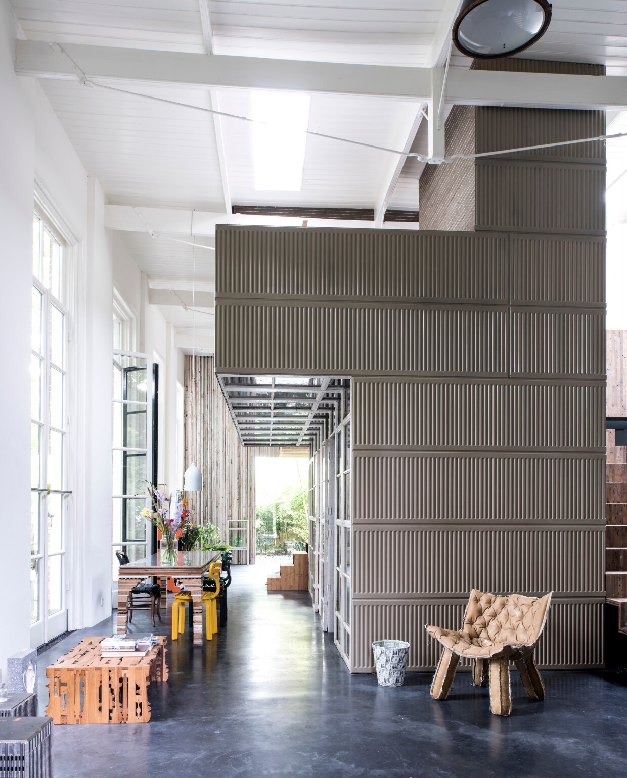 Het vrijstaande object waar onder meer de keuken en slaapkamer in zitten, is aan deze kant opgebouwd uit oude radiatoren uit de barak. De fauteuil is een prototype van Tejo Remy &amp; René Veenhuizen. Daarnaast staat de One Dollar prullenbak van Rolf, die is gemaakt van 150 dollarbiljetten. De tafel maakt Rolf op aanvraag en heet De tafel van het huis. Aan tafel staan Chubby chairs van Dirk vander Kooij. Witte lamp van Studio Wieki Somers. Bloemen van Bloomon. De houten salontafel genaamd Slices of Henry Deux is ontworpen door Rolf. Cut Porcelain van Studio Rolf.fr. De industriële lamp aan het plafond heeft Rolf van Niek Wagemans gekregen. vtwonen 03-2017 | Styling Inge van Lieshout | Fotografie Louis Lemaire/Insidehomepage