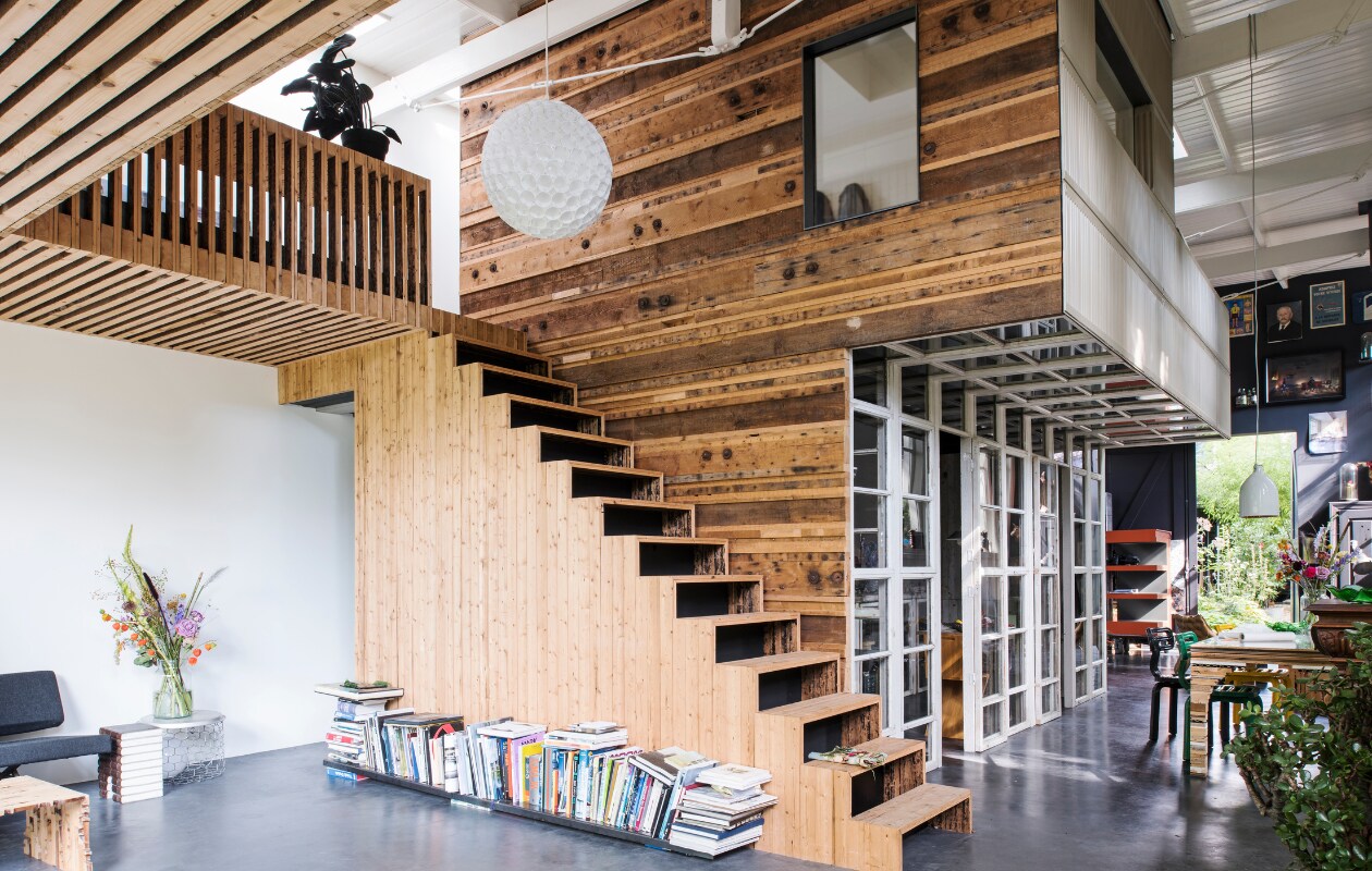 Boven zit de slaapkamer van Rolf en Yffi, daaronder de keuken. Links loop je naar hun walk-in closet. Naast de trap liggen designboeken en tijdschriften. De plant op de eerste verdieping is van vilt en gemaakt door Wandschappen. De witte lamp van plastic bekers is gemaakt door Rolf, maar dit is een bestaand idee. De zwarte bank is van Mart Visser voor Spectrum. Naast de bank staat de Princely Stool, ontworpen door Rolf. Het tafeltje is van Xeen. Mand gekocht in Cambodja. Voor de bank staat een houten salontafel genaamd Slices of Henry Deux, die is ontworpen door Rolf. De wand is opgebouwd uit delen van spanten en de trap met dakbeschot. vtwonen 03-2017 | Styling Inge van Lieshout | Fotografie Louis Lemaire/Insidehomepage