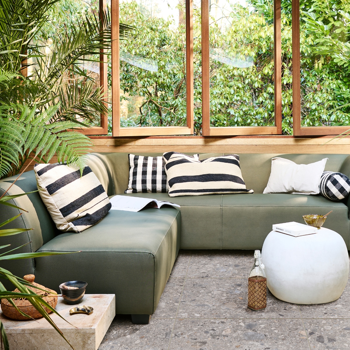 vtwonen collectie (hoekbank Hollandia in camouflage groen, 230x214x70 cm (bxdxh) € 1949,-, ook verkrijgbaar in carbon grijs, als 3-zitter en als 2-zitter, fiberclay bijzettafel Pebble in betonlook wit, 50x35 cm (Øxh) € 179,-, ook verkrijgbaar in 65x49x31 cm (bxdxh) en antraciet, tuintegel Solostone Composite Mix, 70x70 cm per m² € 79,-). M.m.v. vtwonen.nl (gestreepte kussens, waterglas), aimelie.nl (geruite kussens), householdhardware.nl (Tamegroute schaal). Overig privébezit. vtwonen 04-2023 | productie Tanja Saarberg | styling Marlies Does | fotografie Sjoerd Eickmans