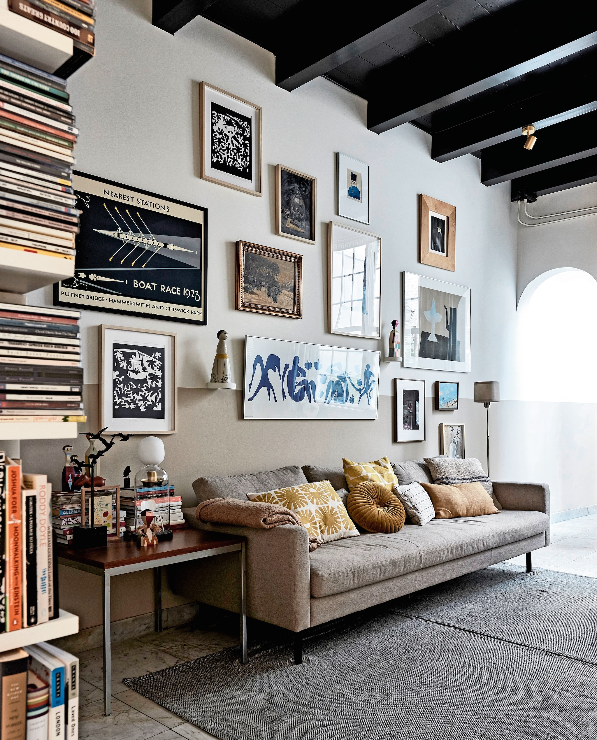 Bank Loods 5. Plaid, rond kussen en effen geel kussen Zara Home. Kussen met zonnetjes, geel-wit kussen, klein kunstwerk op bijzettafel cadeaus. Geblokt kussen zelfgemaakt van Habitat-stof, grijs kussen gemaakt met stof van de markt. Bijzettafel kringloop. Zwart kunstwerk vogels en kleine zwarte vogel familiestukken. Houten beeldjes en leren nijlpaard Vitra. Stolp met model van Revolt-stoel vintage. Tafellamp, vloerkleed en kap stalamp Ikea. Aan de muur: zwart-witte zeefdrukken, schilderij stilleven, persoon met zwarte hoed, groot rechthoekig wit werk in het midden en klein werk vaas met bloemen zelfgemaakt door Heleen. Boat Race 1923 en klein lichtblauw kunstwerk cadeaus. Zeefdruk theepot Klaas Gubbels, ingelijste poster Matisse Ikea, foto Frederique Masselink-van Rijn. Overige makers onbekend. Beeldje gekocht op Bali. Object Vitra. Muurverf  Ral 9010 van Sikkens. Verf lambrisering Pure Mild Sand en plafond Royal Blue van Flexa. Overig onbekend. Beeld vtwonen 02-2024 | styling Fietje Bruijn | fotografie Alexander van Berge