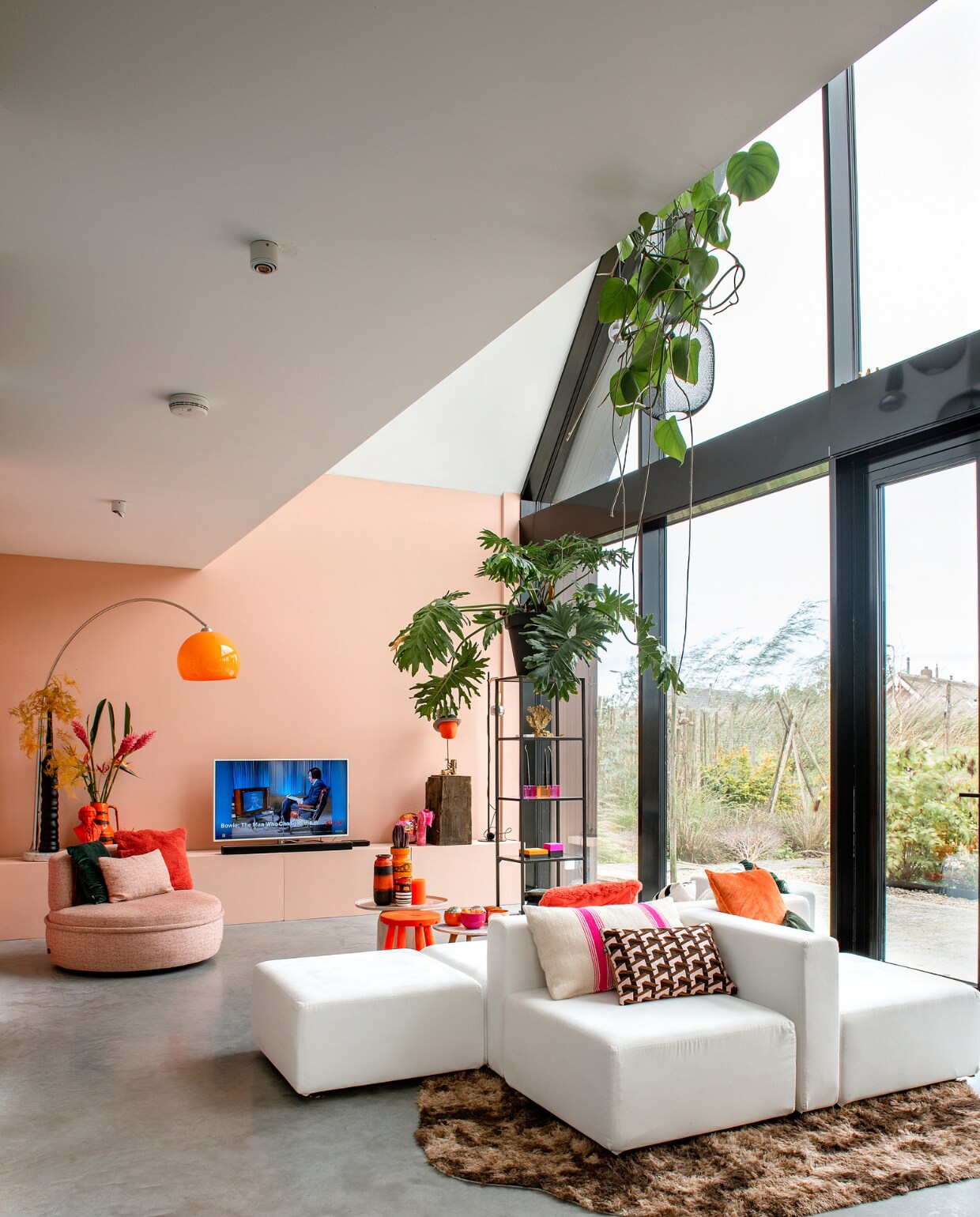 Bank Moduplus, roze bijzettafel Karwei. Kussens en houten blok Nijhof. Stoel Lodge Living, vitrinekast Ikea, vacht Vachten van Terschelling. Roze en oranje kommen en planten De Kaardebol. Oranje hoofd met hoed en oranje vaas links Eigenaar 8. Lamp en aardewerken vazen vintage. Gietvloer Willem Designvloeren, muurverf Blossom Pink van Le Noir &amp; Blanc. Overig onbekend. vtwonen 10-2023 | fotografie Ton Bouwer