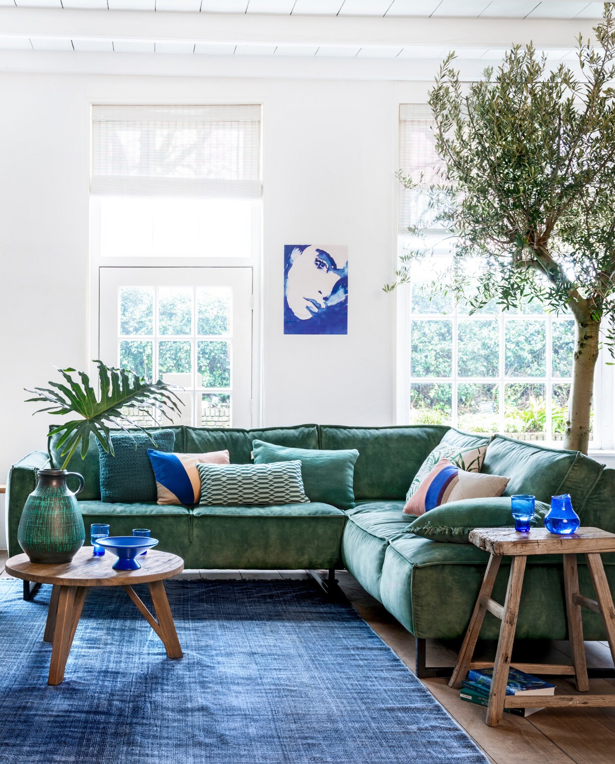Hoekbank Kick, grote salontafel, kleine salontafel, rotan fauteuil Egg Chair (hetkabinet.nl). Gestreept kussen op fauteuil (sissy-boy.com). Groene linnenmix plaid (hm.com).Vloerkleed Vietnam Days, Stonewashed Blue Cotton Block (aaimadewithlove.nl). Boompjes: olijfboom en Eucalyptus (kwekerij). Naturel-groene mand (manggisliving.nl) met plant Schefflera Elegantissima (tuincentrum). Blauw houten gelakte plantenpot (hetkabinet.nl) met plant Kalanchoë (tuincentrum). Blauwe glazen karaf per 3, blauwe fruitschaal van keramiek, hoge blauwe glazen, lage glazen (hkliving.nl). Grote vintage vaas (mippies.com). Kussens op de bank van links naar rechts; vlakken multicolour (hkliving.nl), langwerpig groen gedessineerd (sissy-boy.com), groen stonewashed kelim (hetkabinet.nl), met bladprint (hm.com), met vlakken Patch (hkliving.nl), groen stonewashed Kelim (hetkabinet.nl). Poster (hkliving.nl). Bankje, grote plantenpot links, houten kruk en boeken privébezit. vtwonen 05-2019 | styling Moniek Visser | fotografie Stan Koolen