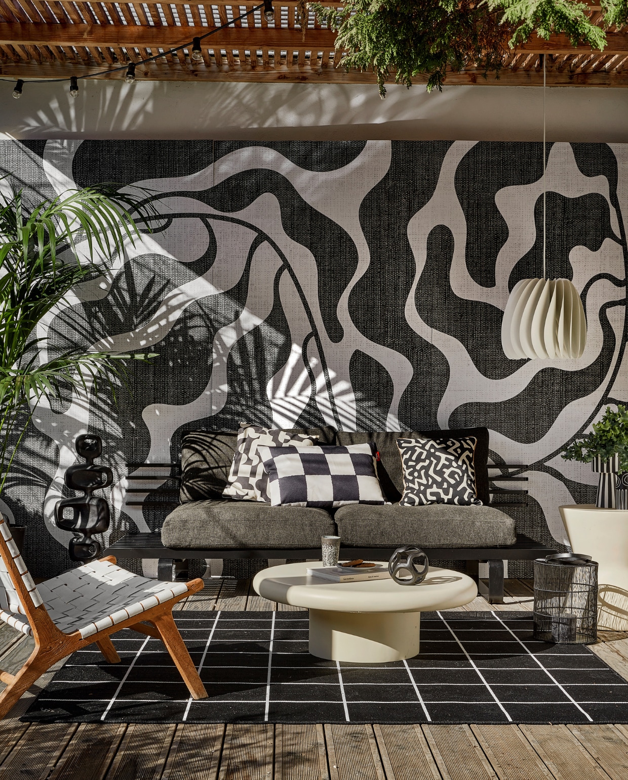 Behang River black & white van Wrap the Wall, per m2 € 150,-, minimale afname 4 m2 (behangfabriek.nl). Zwarte aluminium buitenbank € 1339,80, antraciet tuinkussens, per set € 675,75, zwarte aluminium sculptuur € 295,-, mok 70s, per 2 € 19,99 (hkliving.com). Buitenvloerkleed Svallerup, 200x200 cm € 79,95, hanglamp Skymningen € 79,95 (ikea.nl). Ivoorkleurige polyester bijzettafel, 85x30 cm (Øxh) € 599,-, bijzettafel Pot-Table in Ivory-Slim € 395,- (householdhardware.nl). Zwart-wit kussen Cutro € 10,- (kwantum.nl). Zwart-wit geblokt kussen King Pillow € 55,- (fatboy.com). Kussen Nara € 59,90 (ateliersem.nl). Zwart-wit gestreepte pot van Oyoy € 69,95, aardewerken vaas Troy van Bloomingville € 12,95 (livvlifestyle.nl). Zwart aluminium bolvormig ornament € 31,95, lantaarn € 96,95 (ptmd.nl). Fauteuil met gevlochten zitting privébezit, soortgelijke bij Kave Home (kavehome.com). vtwonen 06-2023 | styling Kim Timmerman | fotografie Peggy Janssen