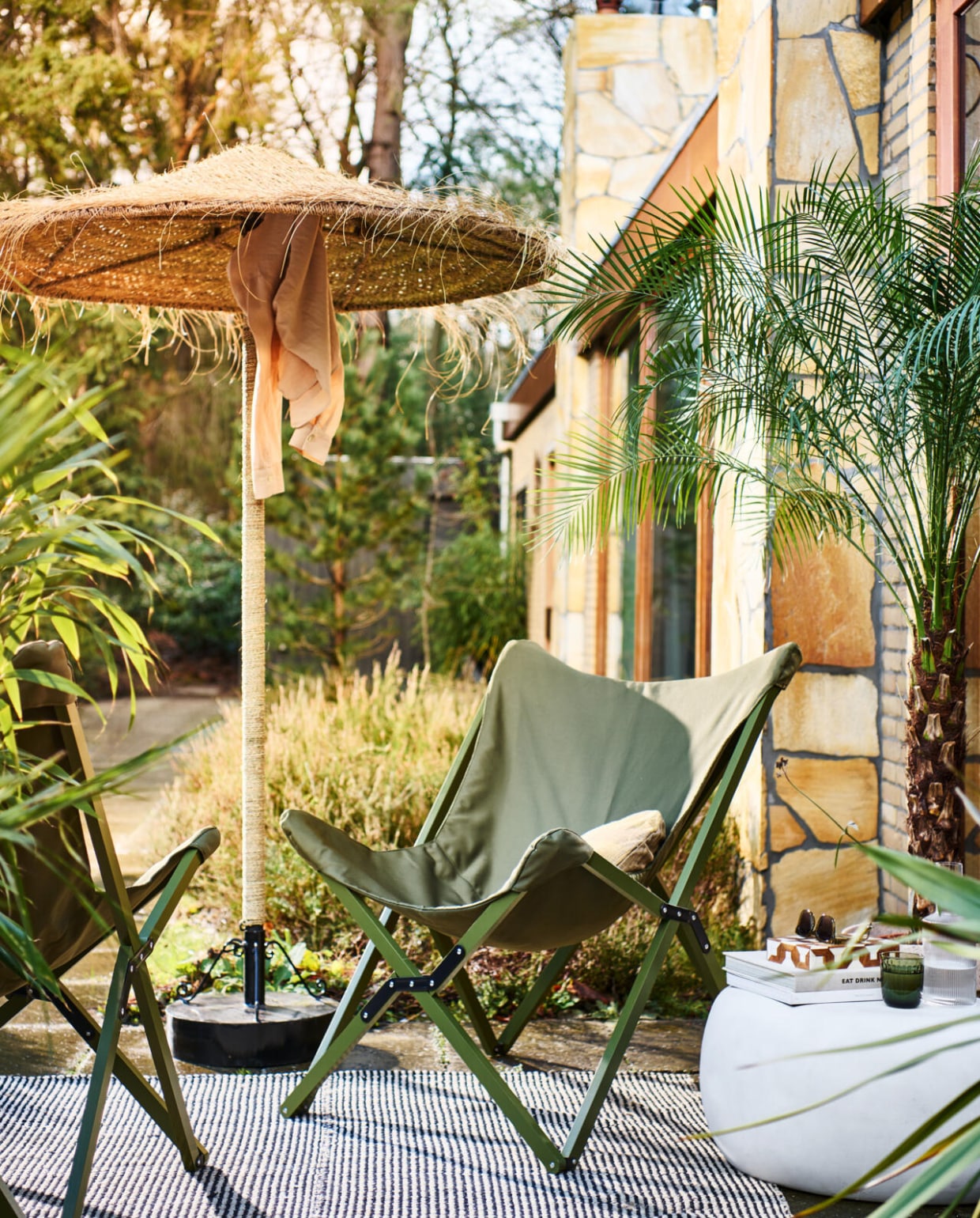 vtwonen collectie (ppvouwbare buitenstoelen Lazy Afternoon in camouflage groen, 85x77x92 cm (bxdxh) € 169,-, fiberclay bijzettafel Pebble in betonlook wit, 65x49x31 cm (bxdxh) € 199,-, ook verkrijgbaar in 50x35 cm (Øxh) € 179,- en in antraciet). M.m.v. householdhardware.nl (parasol, parasolvoet), vtwonen.nl (kussen, waterglas). Overig privébezit. vtwonen 04-2023 | styling Marlies Does | fotografie Sjoerd Eickmans
