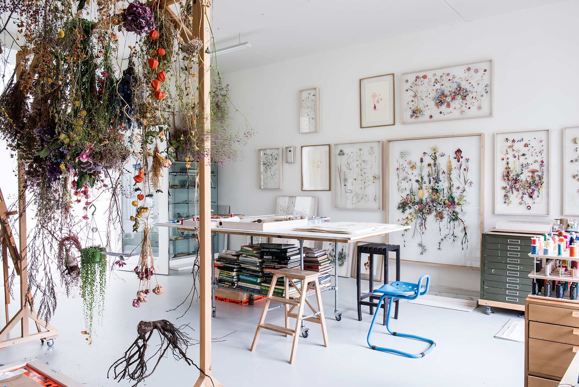 Anne: De meeste bloemen die ik gebruik, komen uit mijn moestuin of de natuur en heel af en toe koop ik bloemen. Ik hang ze te drogen in mijn studio aan een rek dat Vladi voor mij heeft ontworpen. De bloemenpers is ook een ontwerp van Vladi. De lijsten zijn gemaakt door Vladi, net als de houten kist om werk in te versturen. Schildersezel gekocht op Marktplaats. Haar bureau van Marktplaats. Het kastje is ontworpen door Vladi vtwonen 07-2017 | Fotografie Louis Lemaire/Inside Homepage | Styling Esther Jostmeijer