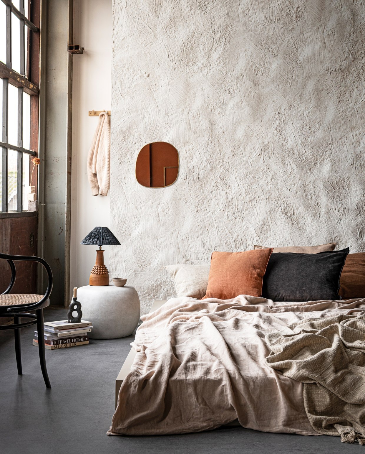 Muurverf in Chalk White (pure-original.nl). Bed op maat gemaakt en geverfd met Farrow &amp; Ball in Oxford Stone (com-anders.nl). Linnen dekbedovertrek in Sand van Linge Particulier, vanaf € 325,-, kussen Sabra offwhite Oblong XL € 113,-, linnen kussen Vice Versa van Maison de Vacances € 133,-, kussen Vice Versa Canvas Formentera van Maison de Vacances € 207,-, kussen Vice Versa Toile Nomade van Maison de Vacances € 161,-, messing spiegel Ame van Caravane € 141,- (couleurlocale.eu). Zwart kussen van vtwonen collectie € 34,95 (vtwonen.nl). Bijzettafel Pebble van vtwonen collectie € 129,-(basiclabel.nl). Lamp van Madam Stoltz € 231,75 (deens.nl). Kandelaar van Malwina Kleparska € 95,-, boek van Bea Mombaers € 43,50 (studio-yen.com). Kom van klei € 9,- (household- hardware.nl). Kapstok van ByOn € 24,95 (littledreamers.nl). Stoel en overige boeken privébezit. vtwonen 03 2021 | styling Marlies Does, fotografie Sjoerd Eickmans