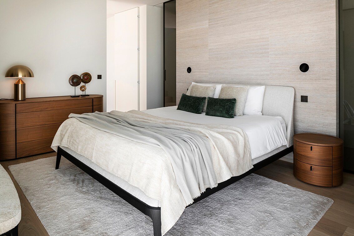 Het gestoffeerde bed Kelly is van Poliform. De commode en het nachtkastje zijn beide van walnoothout en komen uit de lijn Onda van hetzelfde merk. De goudkleurige lamp Atollo is van Oluce. Jonah Samyn