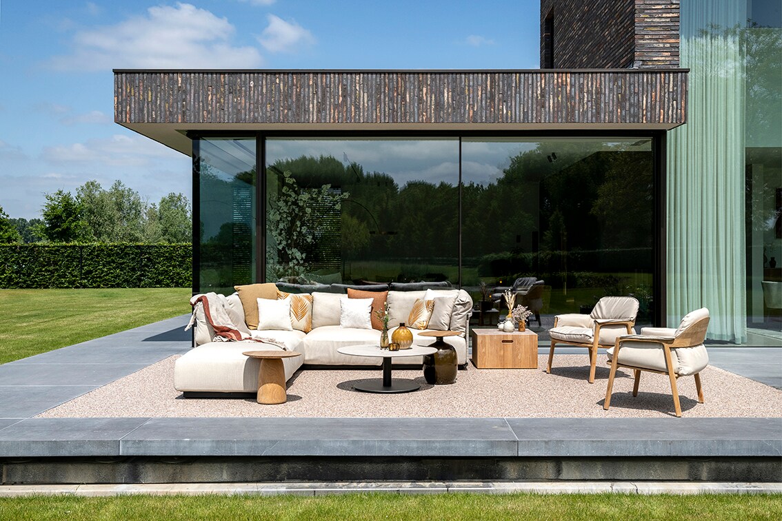 De outdoor loungehoek bestaat uit L-vormige zitbank Nodi en twee loungechairs Nomad. De lage, ronde T-table, de organisch vormgegeven sidetable Gloster Blow, de massief teakhouten sidetable Gloster Block en bijzettafel Dunes maken hem compleet. Alles van Tribu. Jonah Samyn