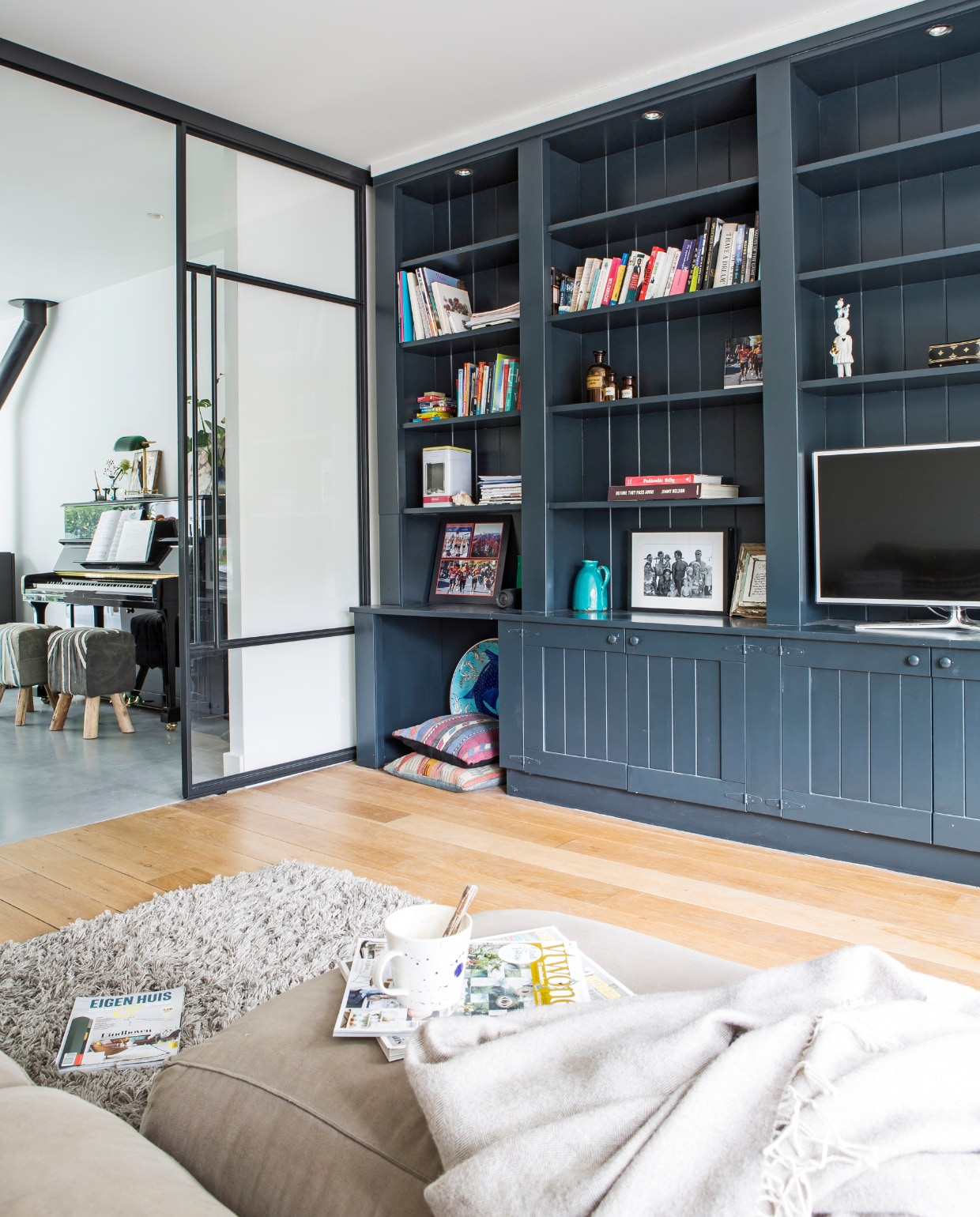 De voorkamer, met zelfbouwkast van Hubo, geverfd in kleur Railings van Farrow &amp; Ball. Op de vloer ligt opgeschuurd hout. De bank is van De Tafelaar, de lichte plaid van Ikea. In de kast een tv van Samsung, een turquoise kruikje van de rommelmarkt en kelimkussens van Loods 5. Het vloerkleed is van Ikea, de glazen deuren met stalen frame zijn gemaakt door Hogendoorn Metaal. De piano komt uit een winkel in het dorp van Leonies ouders, Druten. Stoeltjes ervoor van Tica, lampje erop gevonden op Ebay. vtwonen 12-2017 | fotografie Henny van Belkom