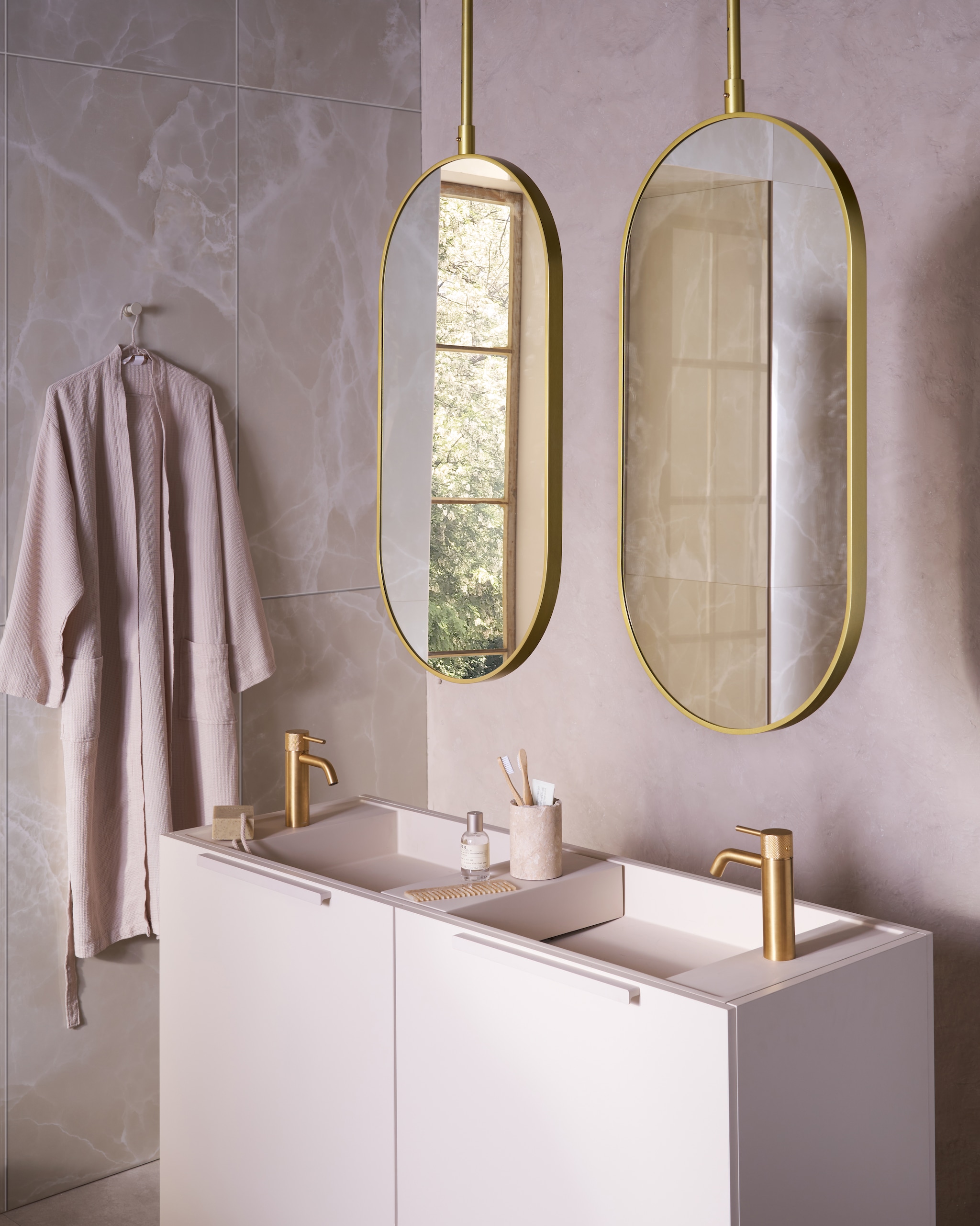 Op de wand tegels Onyx Crystal White, 60x120 cm, per m² € 92,50. Spiegels Oval in goud, 60x90 cm (bxh) € 550,-, ook verkrijgbaar in andere afmetingen. Top Solid onderkast in matzand, 124,6x44,4x90 cm (bxdxh) € 1300,-, Top Solid wastafel Wide in zand, 121x40,5x20 cm (bxdxh) € 1095,-, Top Solid plateau voor wastafel in zand, 36,8x15x8 cm (bxdxh) € 208,-. M.m.v. zarahome.com (beker, zeep, kimono),  plieger.nl (kranen Roma in geborsteld goud), lelabofragrances.com (parfum). vtwonen 09-2024 | styling Cleo Scheulderman | fotografie Jeroen van der Spek